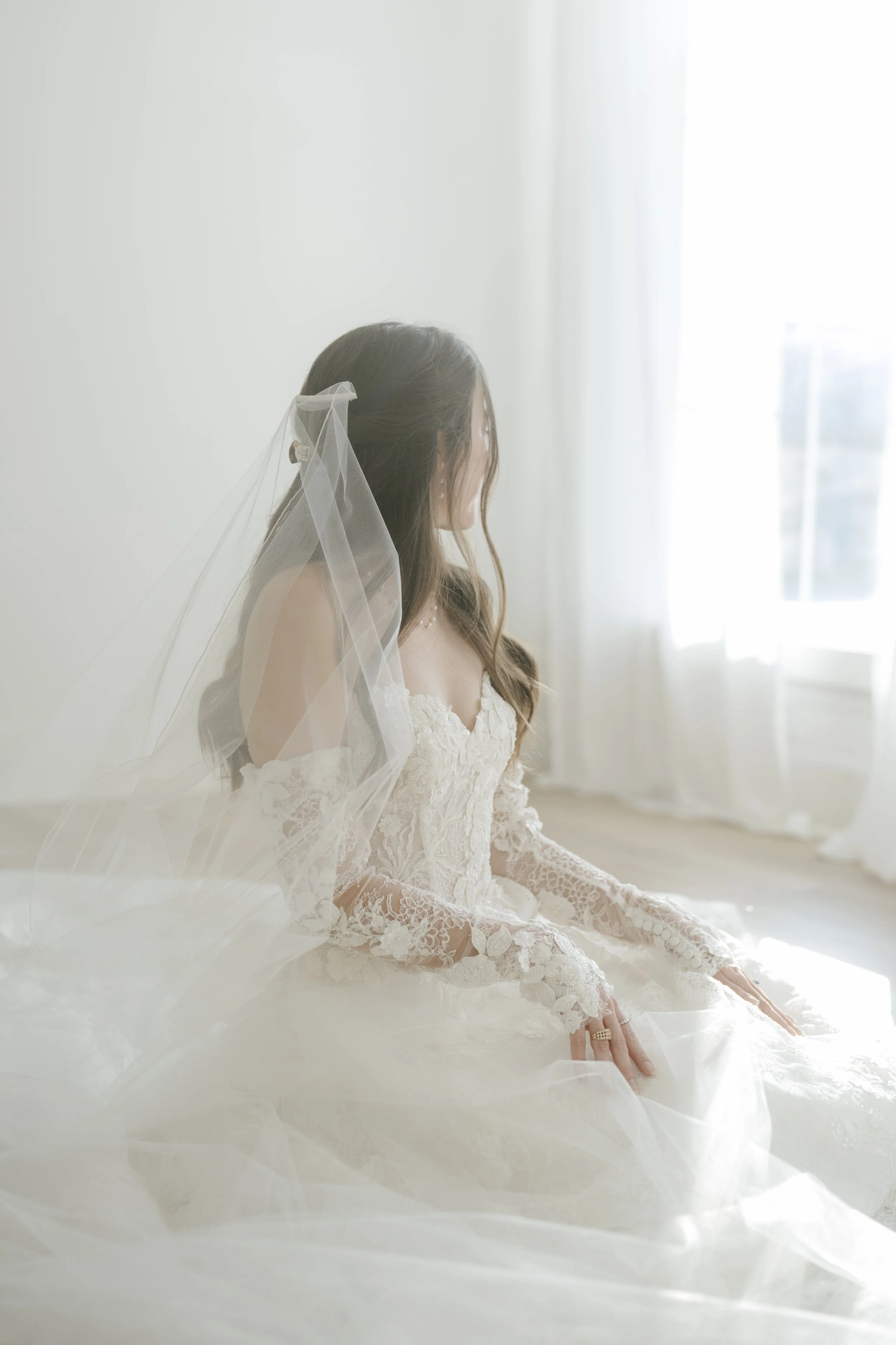 A bride in a lace wedding dress and veil sitting on the floor near a window with white curtains, looking away.