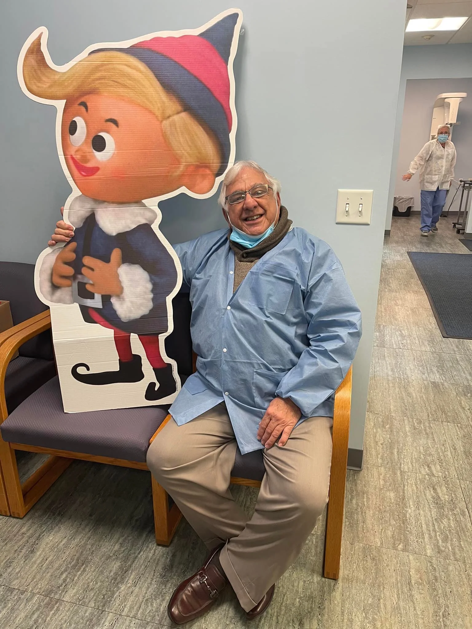 An elderly man sitting on a bench, smiling, holding a large cartoon cutout of a girl with blonde hair, wearing a red and blue striped hat, in a medical or healthcare setting.