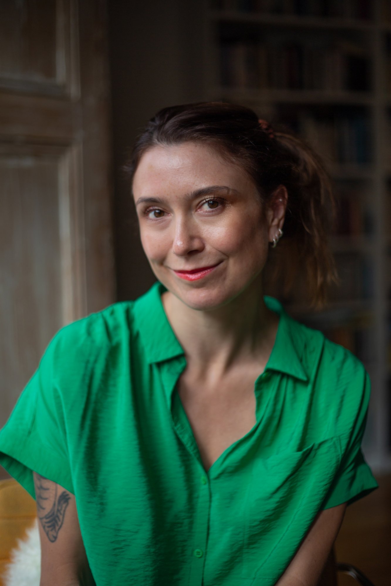 A woman with a tattoo on her left arm wearing a bright green shirt, smiling and looking at the camera inside a room with wooden furniture and bookshelves in the background.