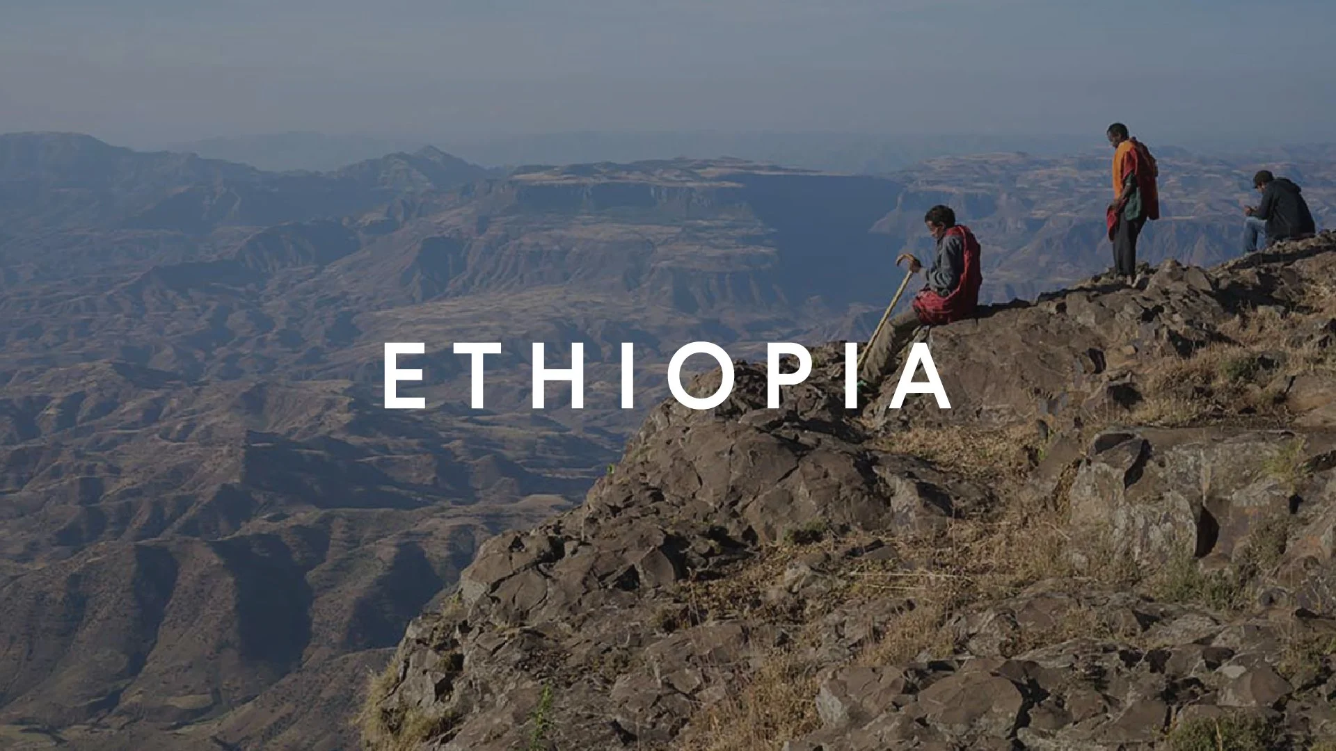 Three hikers sitting and standing on a rocky ledge overlooking a vast mountain landscape in Ethiopia, with the word Ethiopia overlayed in white text.