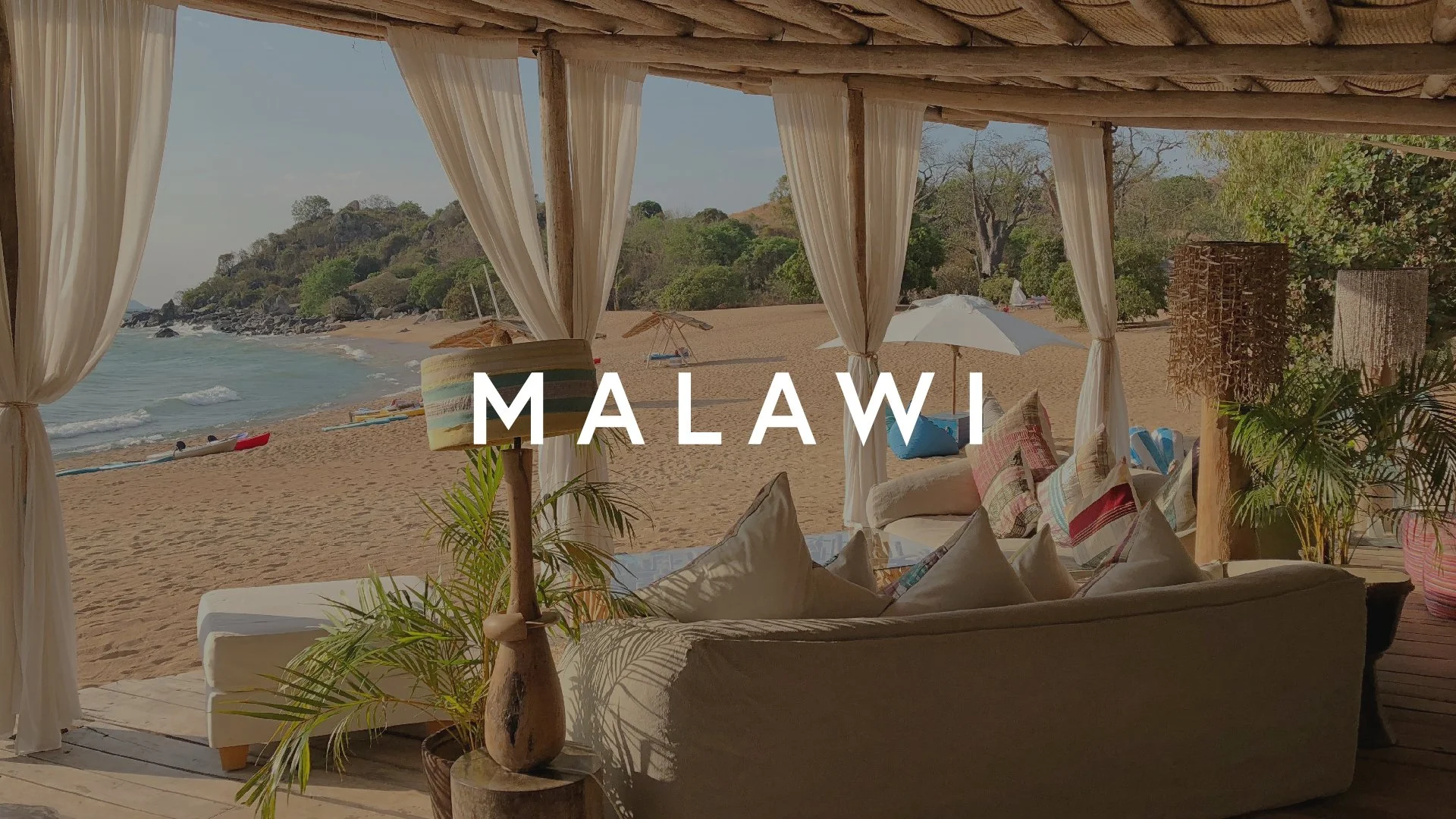 Beachside cabana with white curtains, wooden furniture, and colorful cushions. Beach with umbrellas, rocks, and trees in the background in Malawi.