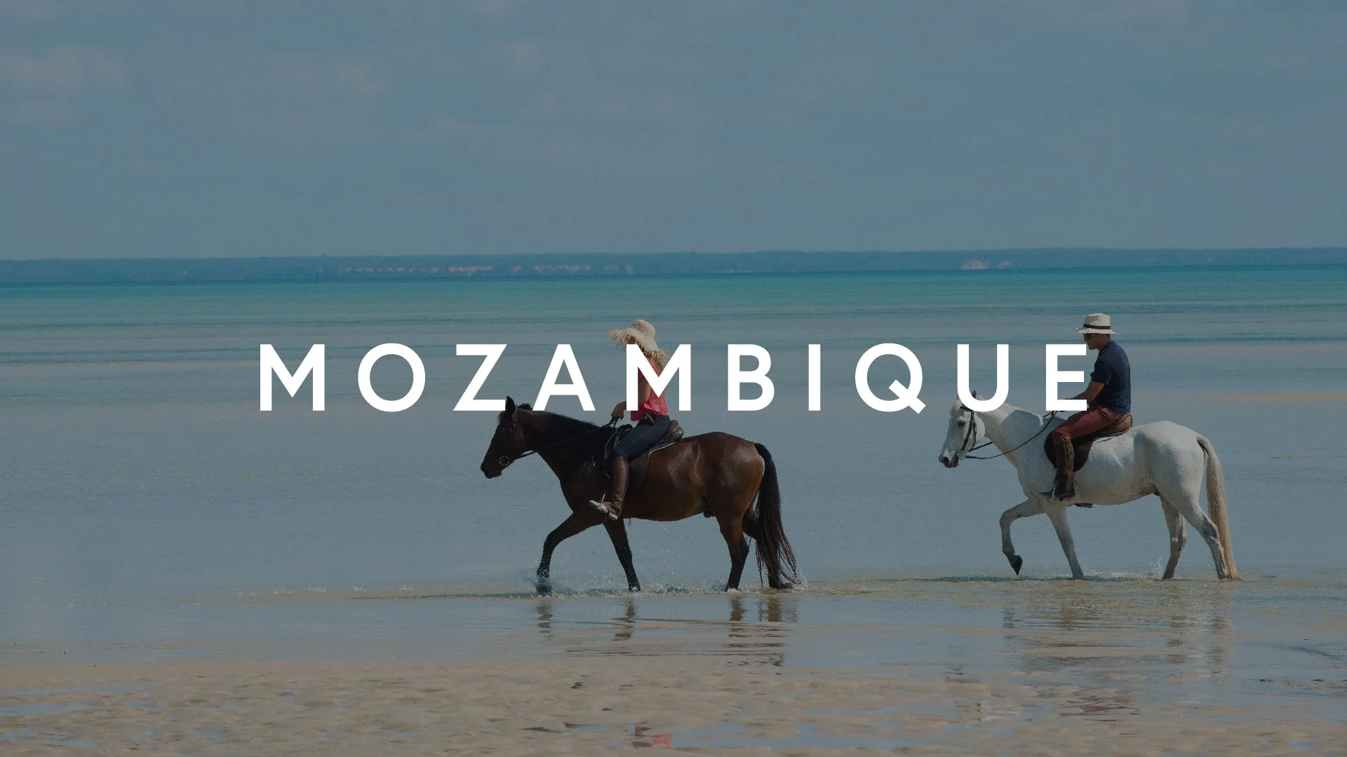 Two people riding horses in shallow water on a beach in Mozambique, with the ocean and sky in the background.