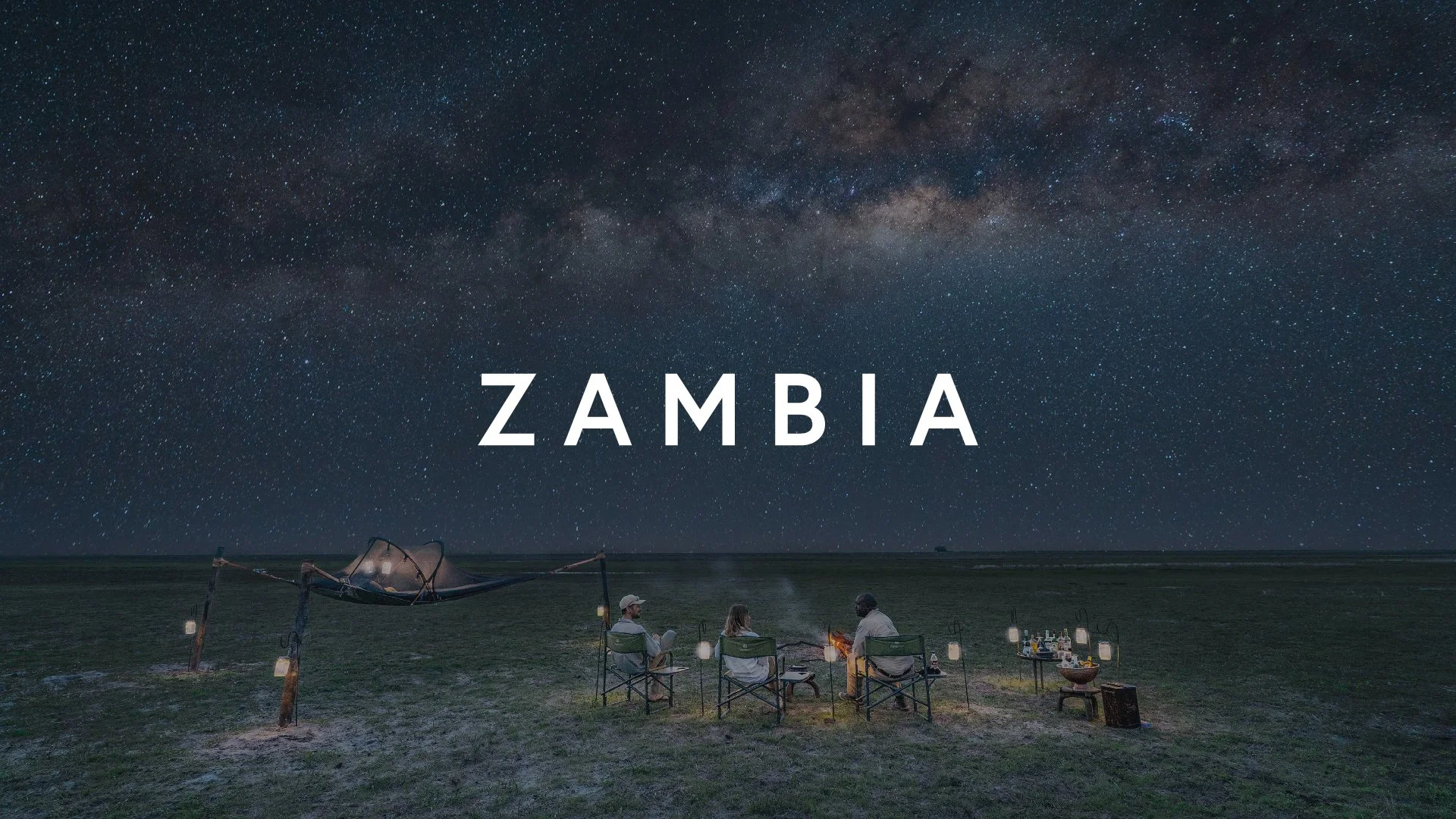 Night sky filled with stars and the Milky Way above a group of people sitting around a campfire on a grassy field in Zambia.
