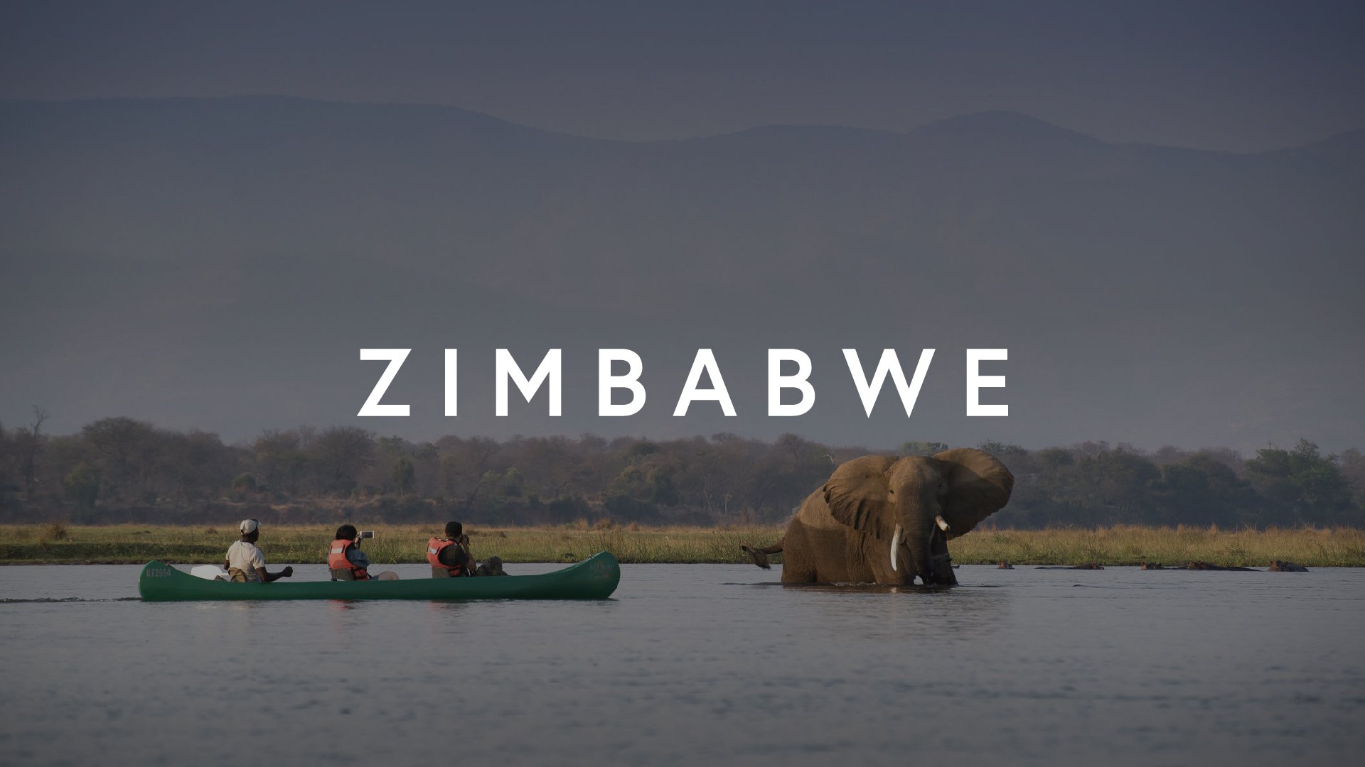 A water scene in Zimbabwe showing a group of three people in a canoe with life jackets, and an elephant standing in the water, with a background of trees and mountains.