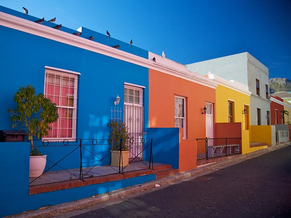Colorful row of houses painted in bright blue, orange, yellow, and teal with potted plants and pigeons on the roof against a clear blue sky.