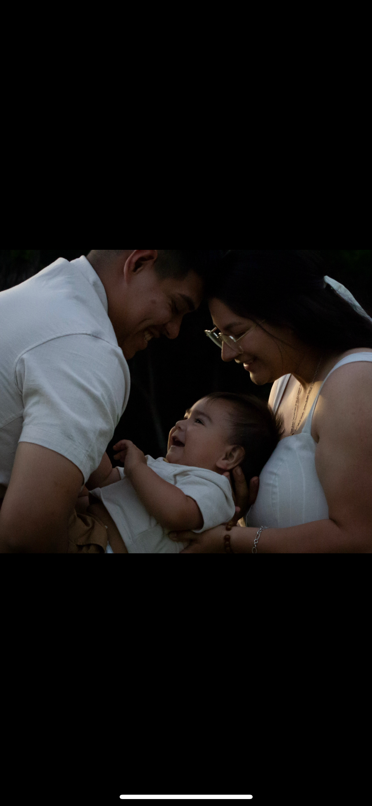 Family of three smiling and playing together outdoors in the evening, with a dark background and warm light on their faces.