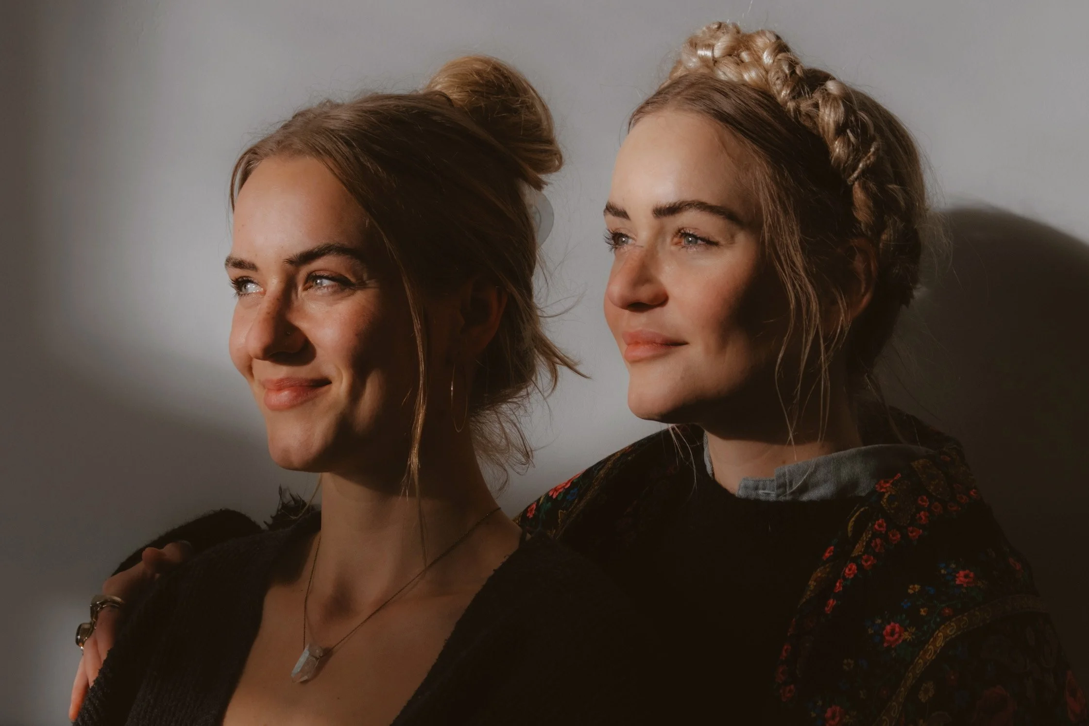 Two women with light brown hair, smiling and standing close together, with one woman having her hand on the other's shoulder against a plain background.