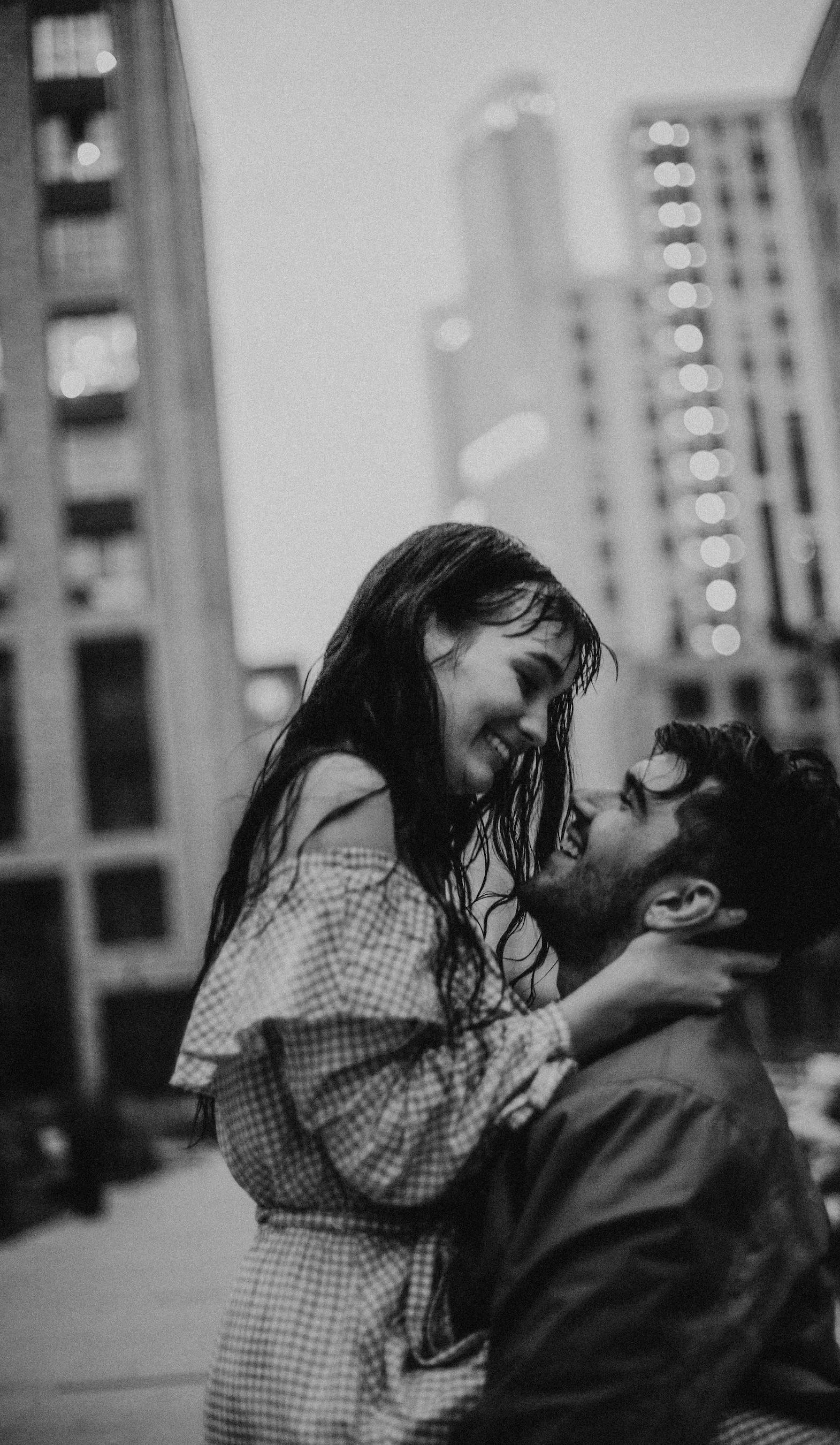 A black and white photo of a happy couple in an urban setting, with buildings in the background. The woman is sitting on the man's lap, both smiling and looking at each other closely.