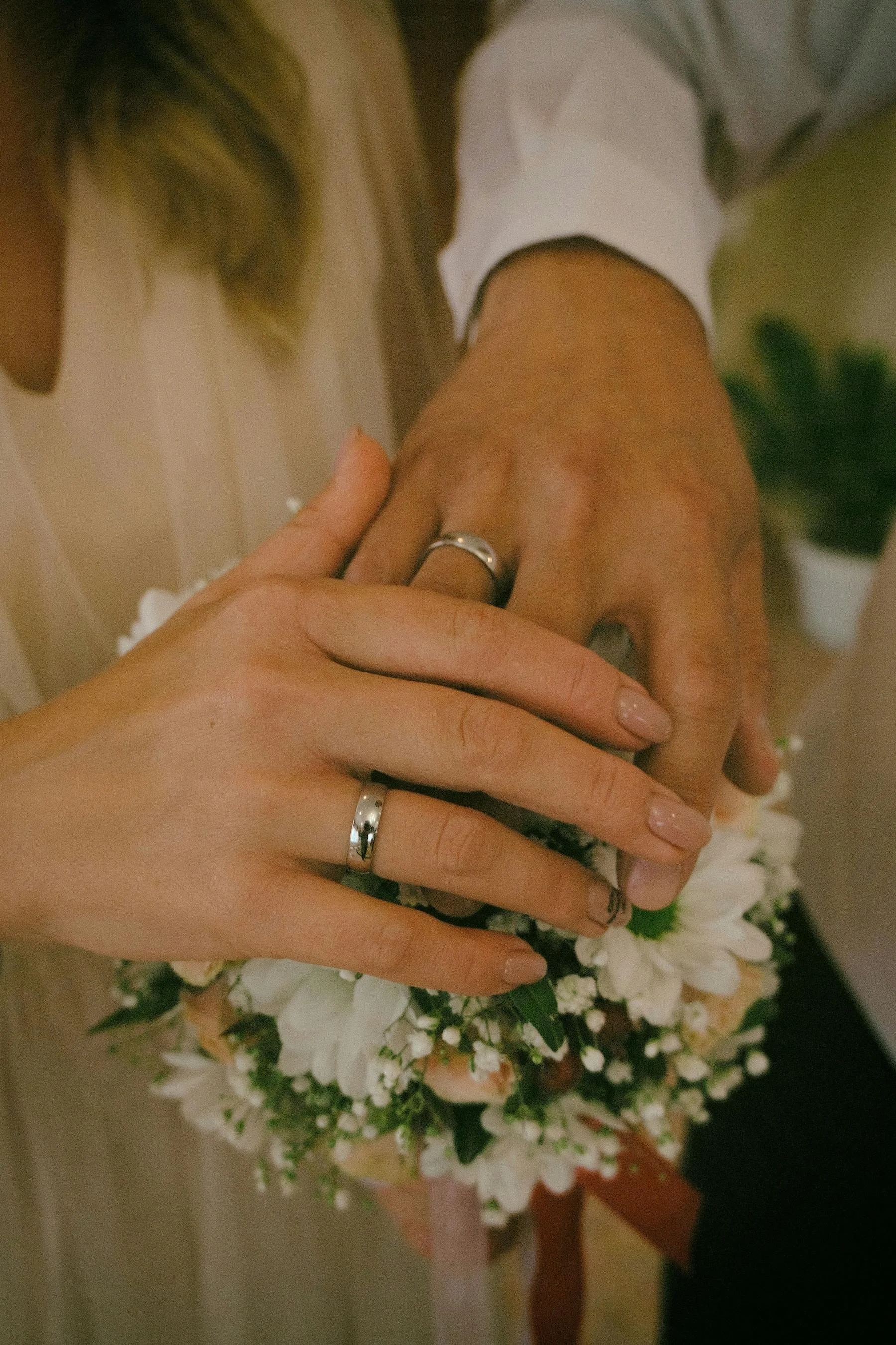 Close-up of a bride and groom's hands showing wedding rings, resting on a bouquet of white flowers.