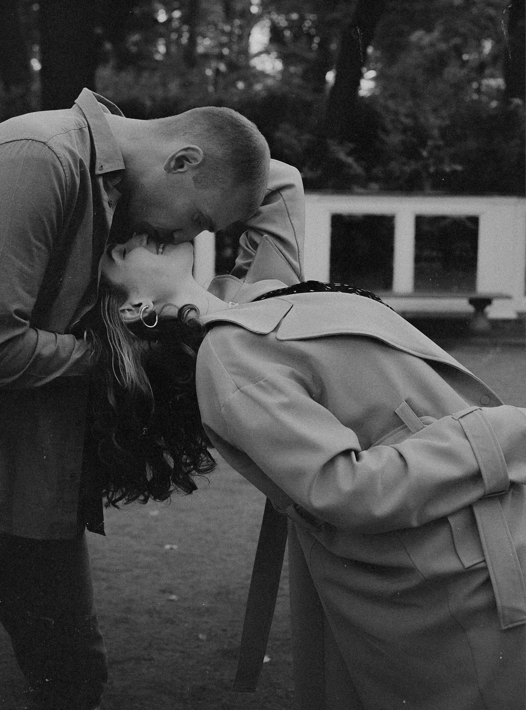 A black-and-white photo of a couple sharing a passionate kiss outdoors, with the man leaning over the woman, who is lying back on a table or bench, in a park-like setting.