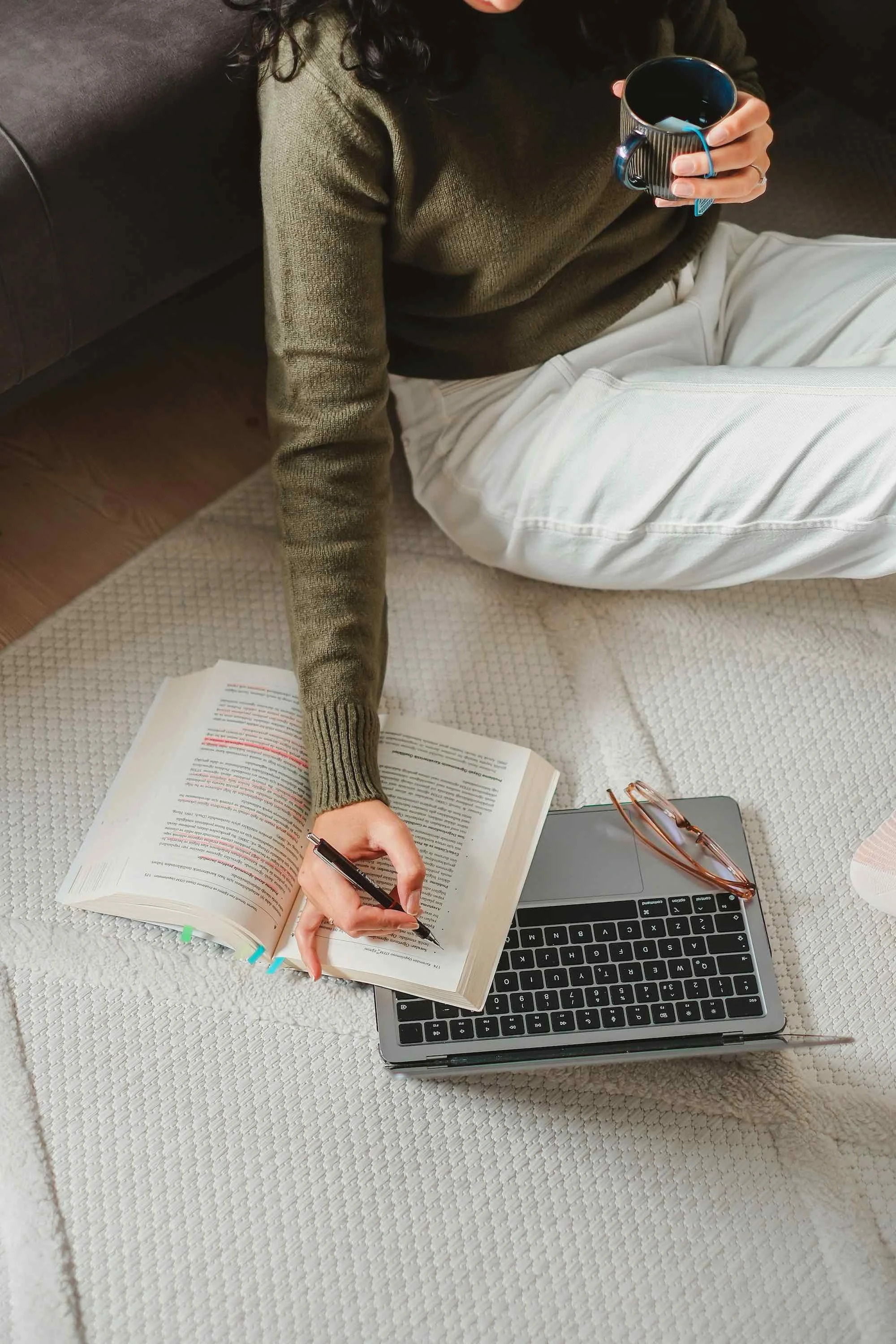 Person sitting on a blanket or rug on the floor, reading a book with highlighter marks, holding a mug, with a laptop and glasses nearby.
