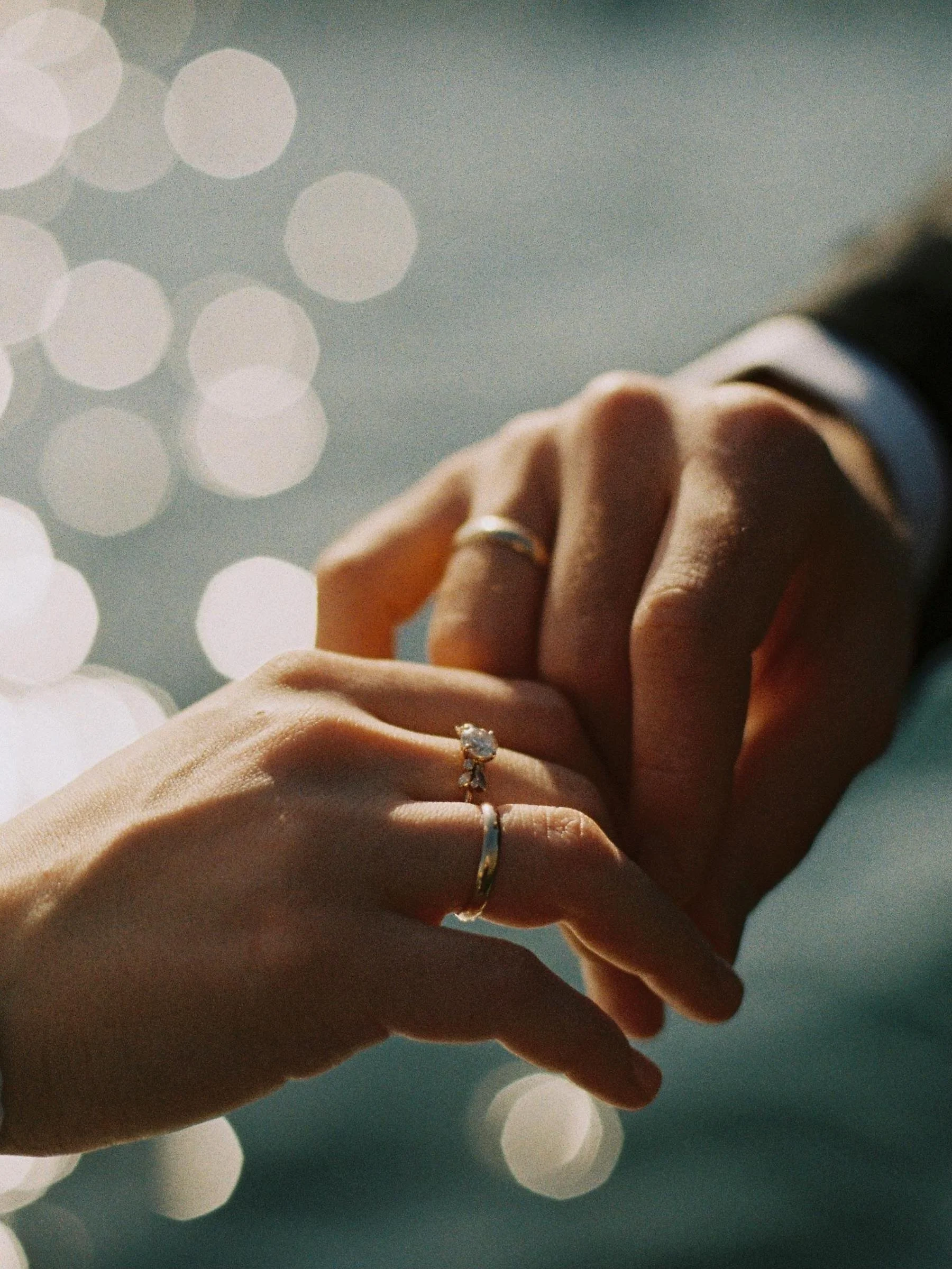 Close-up of hands with wedding rings, holding each other, with blurred bokeh lights in the background.