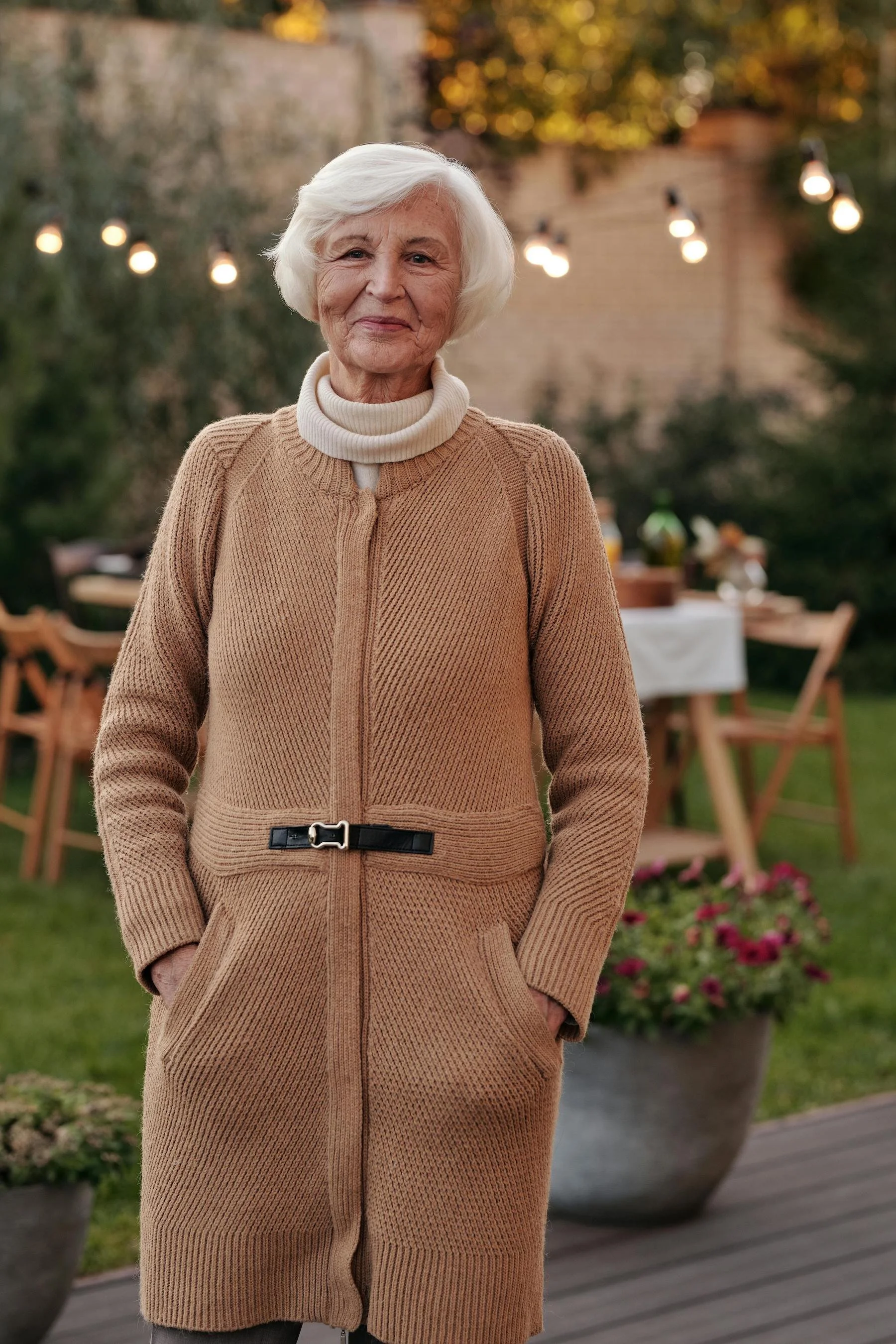 An elderly woman with white hair stands outdoors during evening, dressed in a beige knitted coat with a white turtleneck underneath, smiling softly with a garden and string lights in the background.