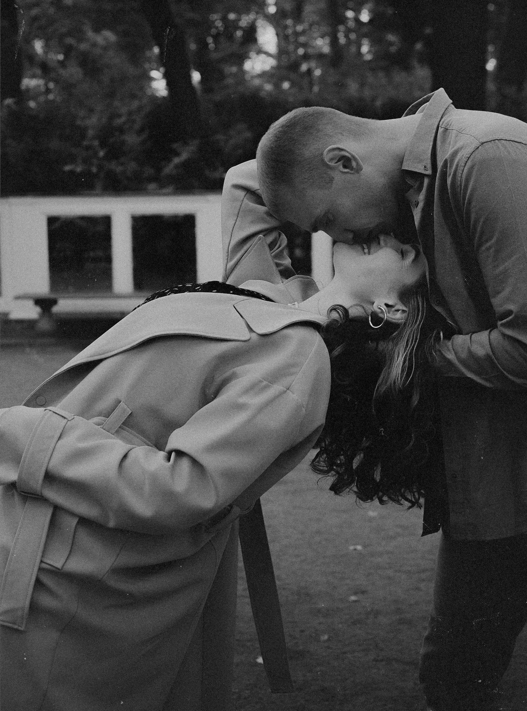 A black-and-white photo of a man and woman sharing a kiss outdoors, with the woman leaning backward and the man leaning forward over a railing.