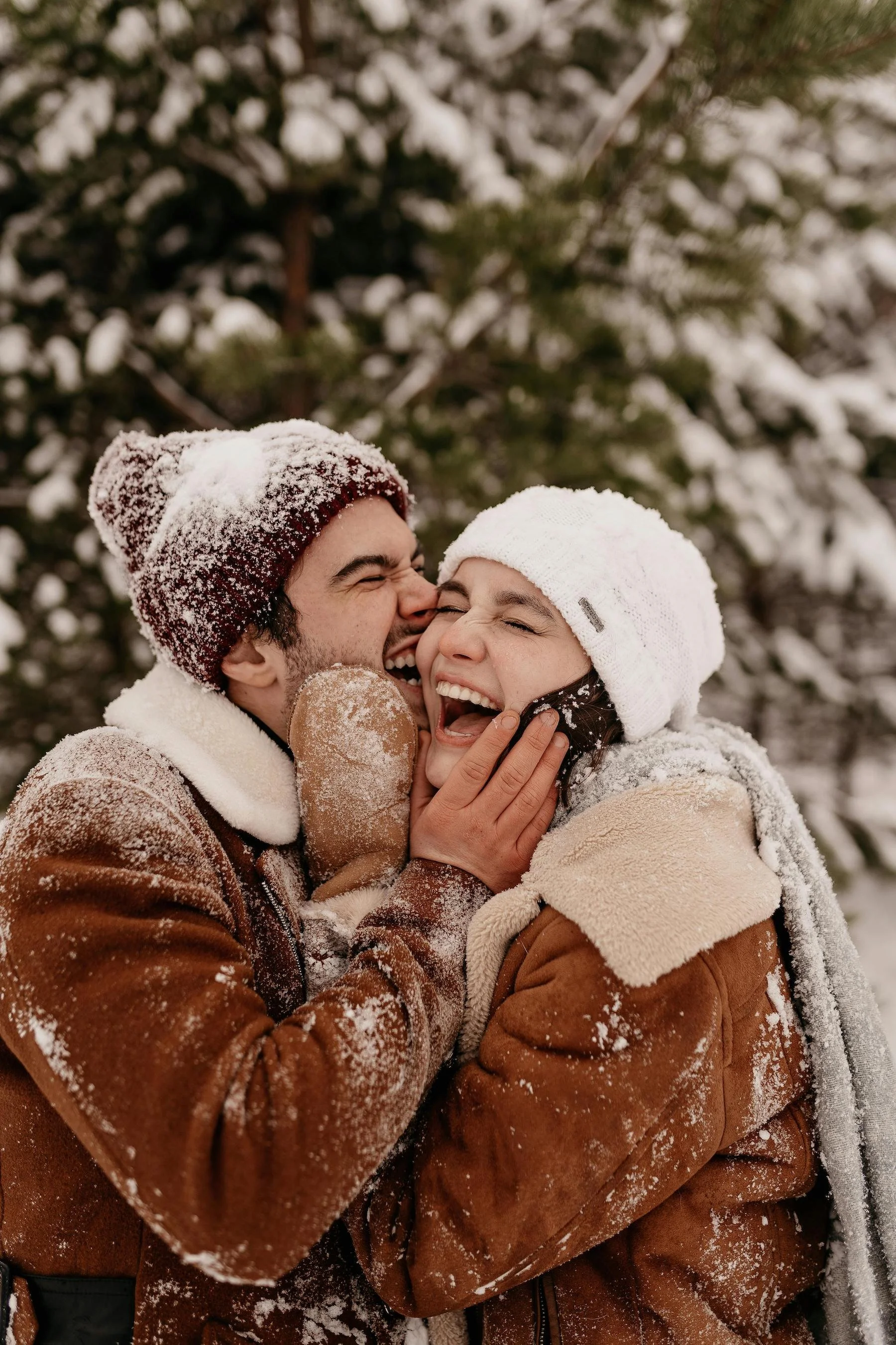 Two people in winter clothing, laughing and embracing each other outdoors in a snowy forest.