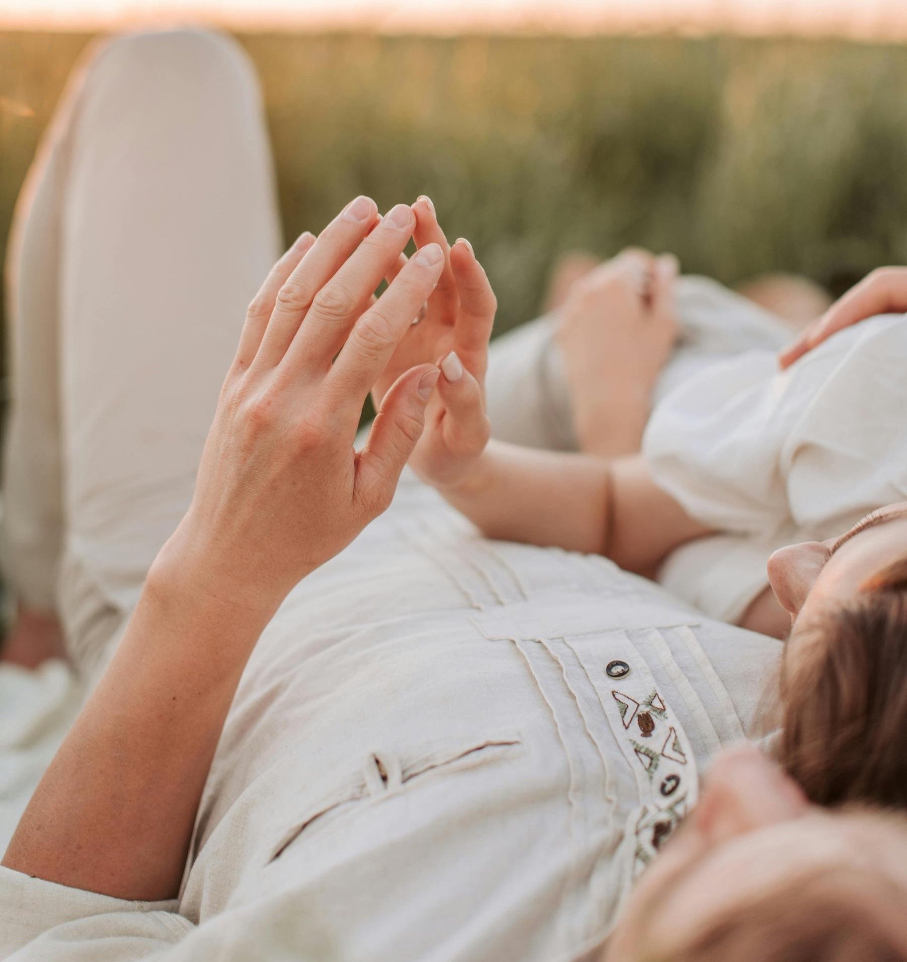 Two women lying on grass, holding hands with palms together in a prayer or meditative gesture, with a sunset or golden hour background.