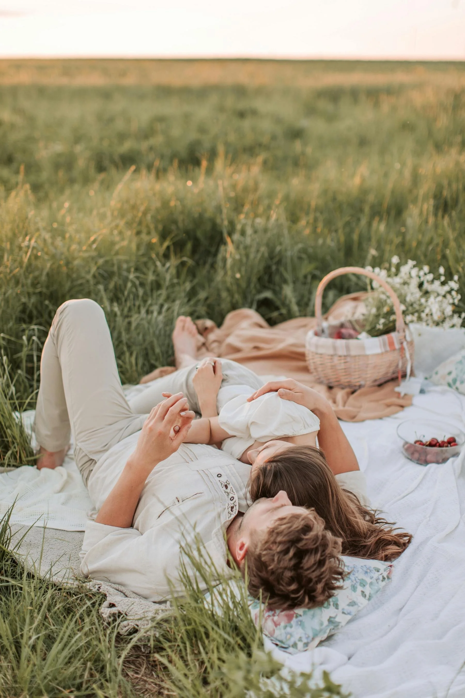 A couple lying on a picnic blanket in a grassy field during sunset, embracing each other, with a basket and bowl of cherries nearby.