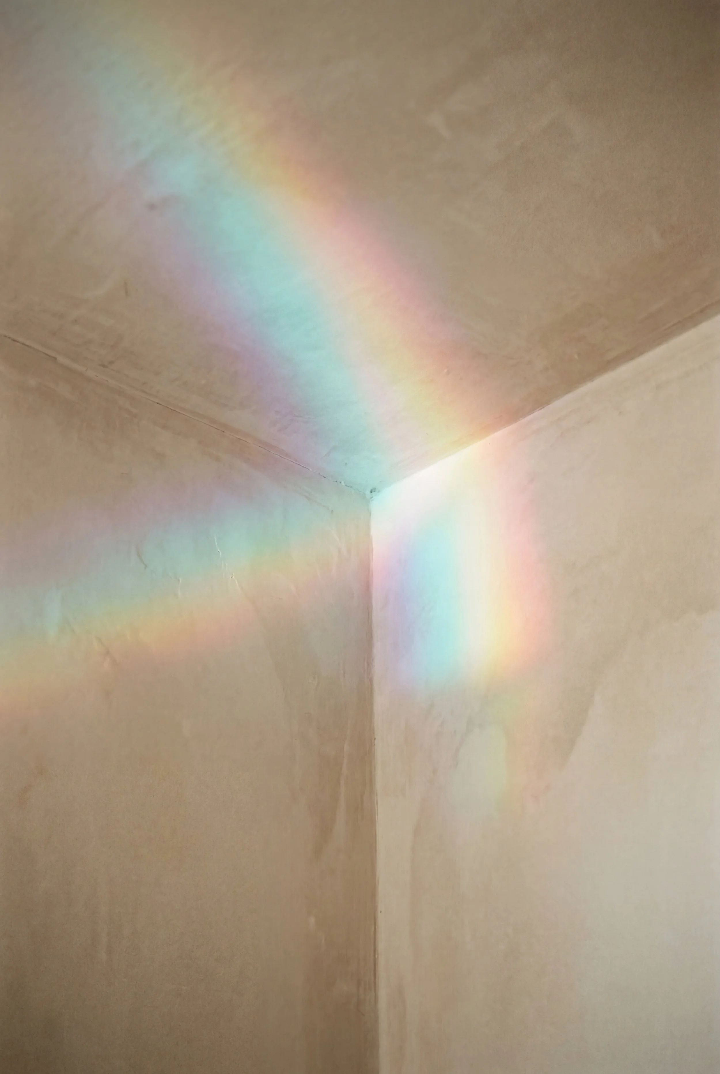 Colorful rainbow-like light reflections on the corner of a beige room, created by sunlight passing through a prism or glass.