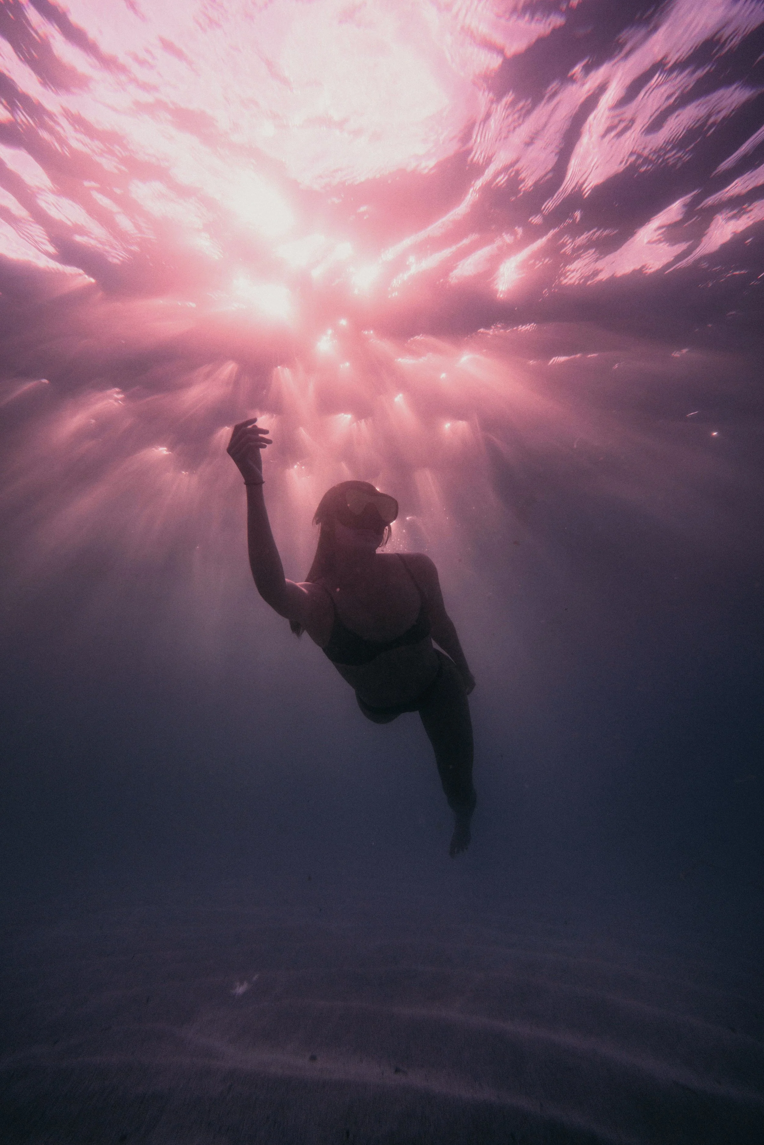 A woman underwater wearing a diving mask, reaching toward the sunlight filtering through the water.