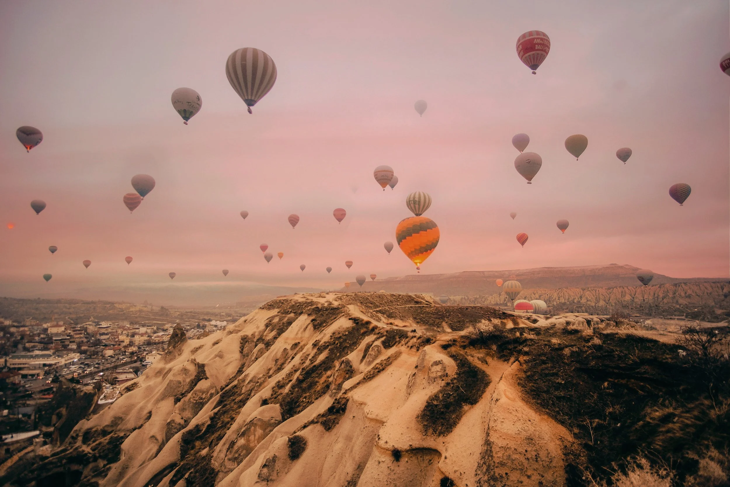 Multiple hot air balloons floating above a rocky landscape at sunrise or sunset, with a city visible in the distance.