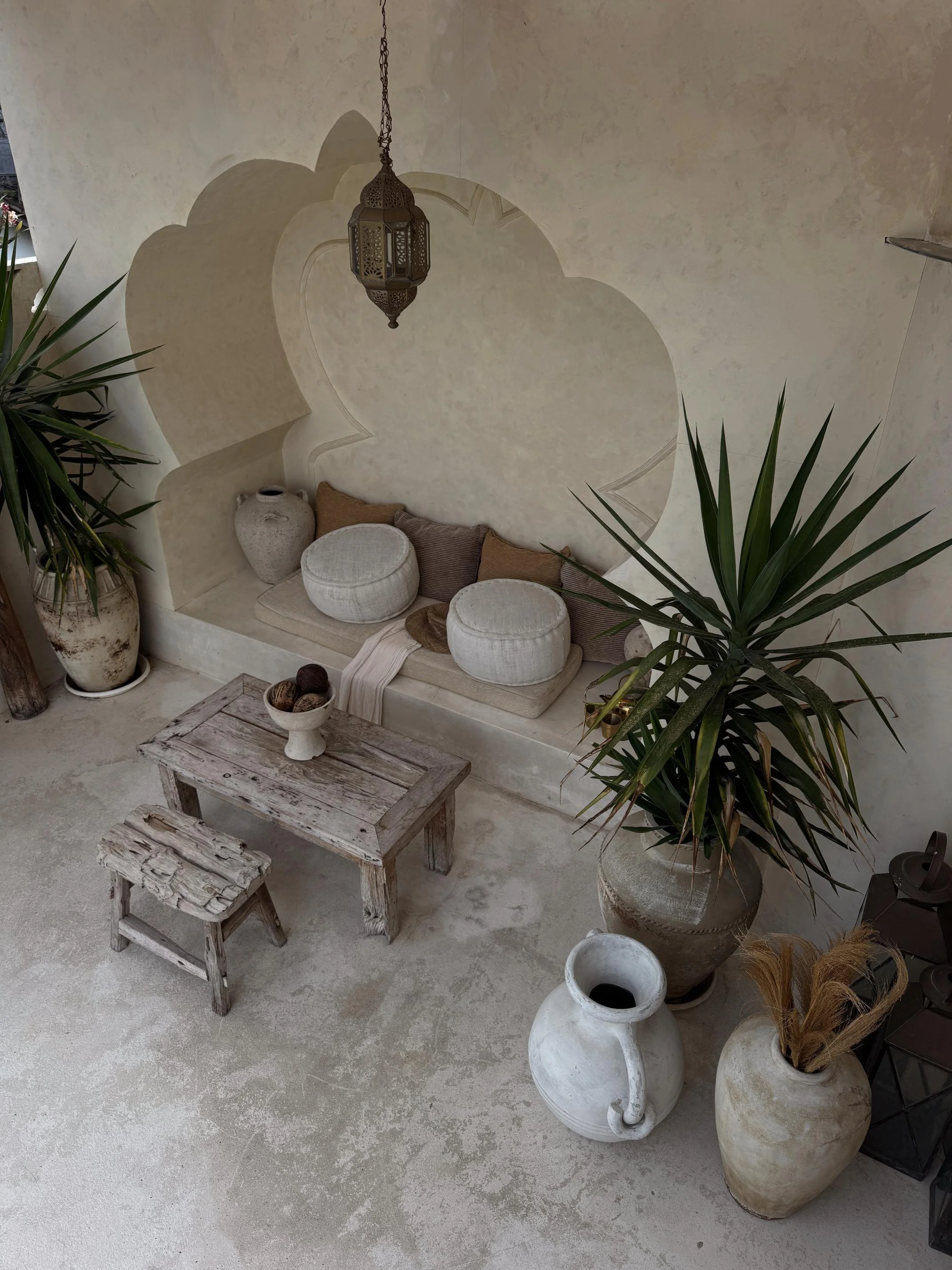 Bohemian-style living space with a built-in nook seating area, a wooden coffee table, two large potted plants, and decorative vases, with a hanging lantern overhead.