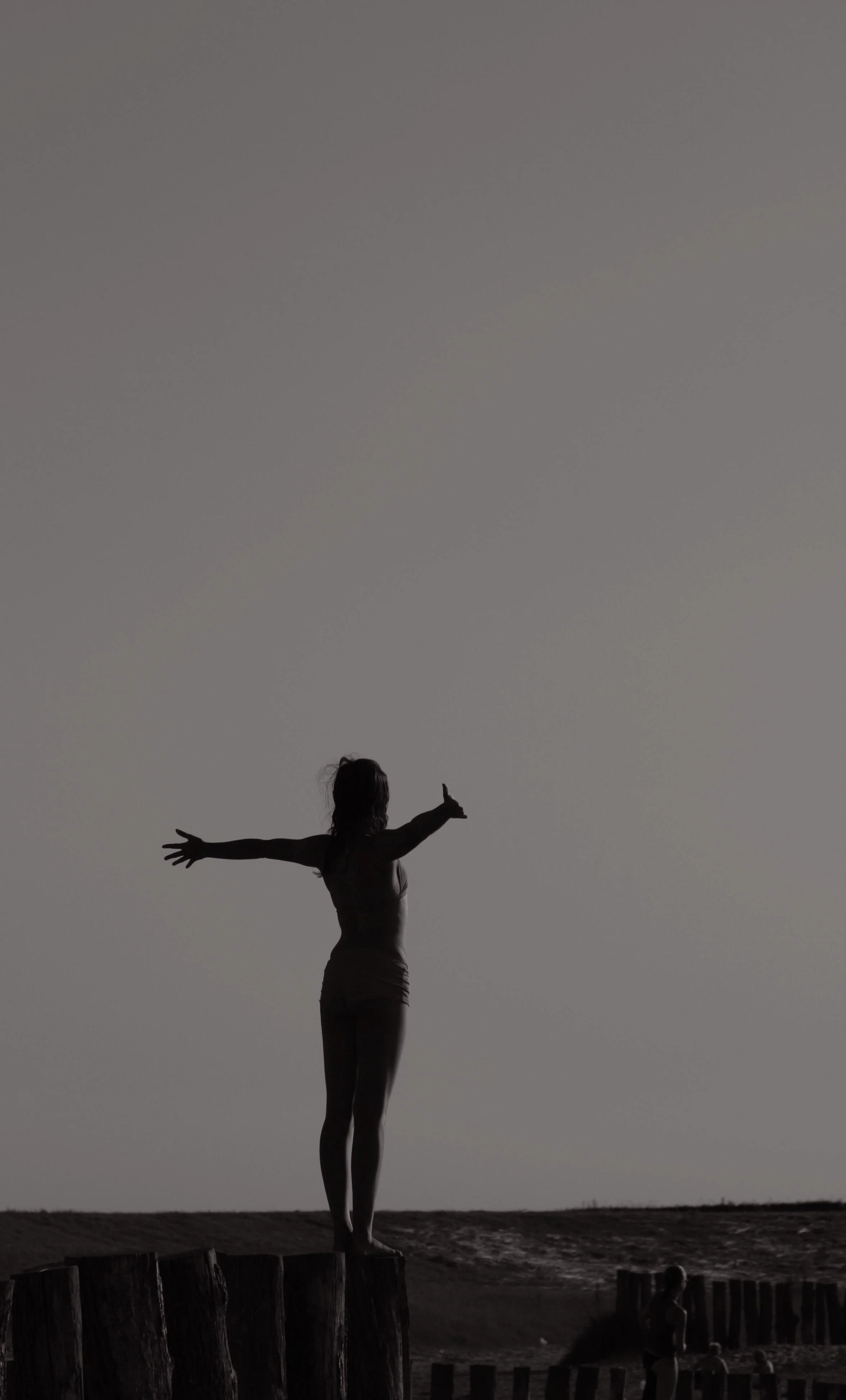 A silhouette of a woman standing on a wooden post with arms outstretched and thumbs up at the beach, with the ocean and sky in the background.