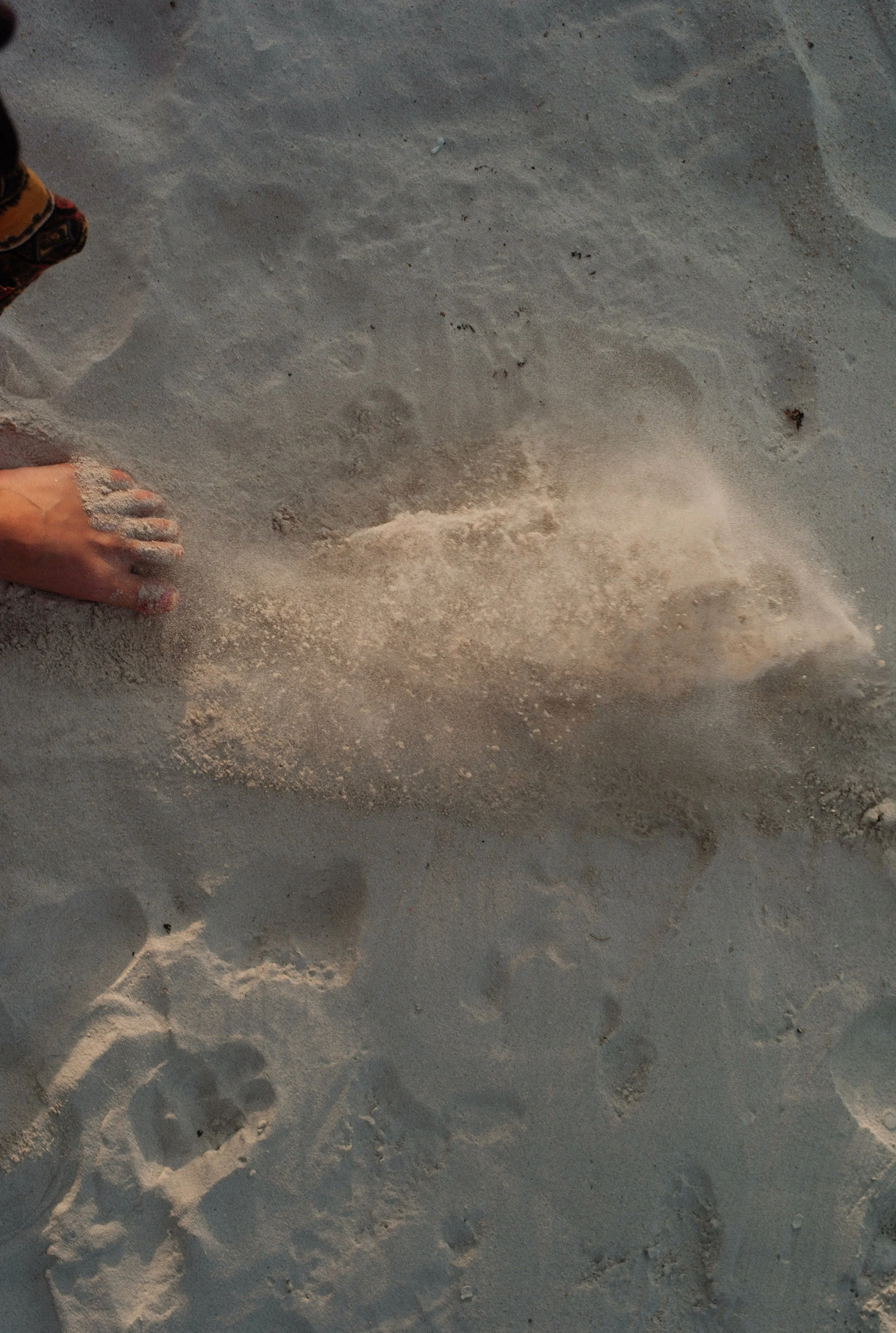 A person using their foot to kick a clump of sand on a sandy beach, leaving a puff of sand in the air.