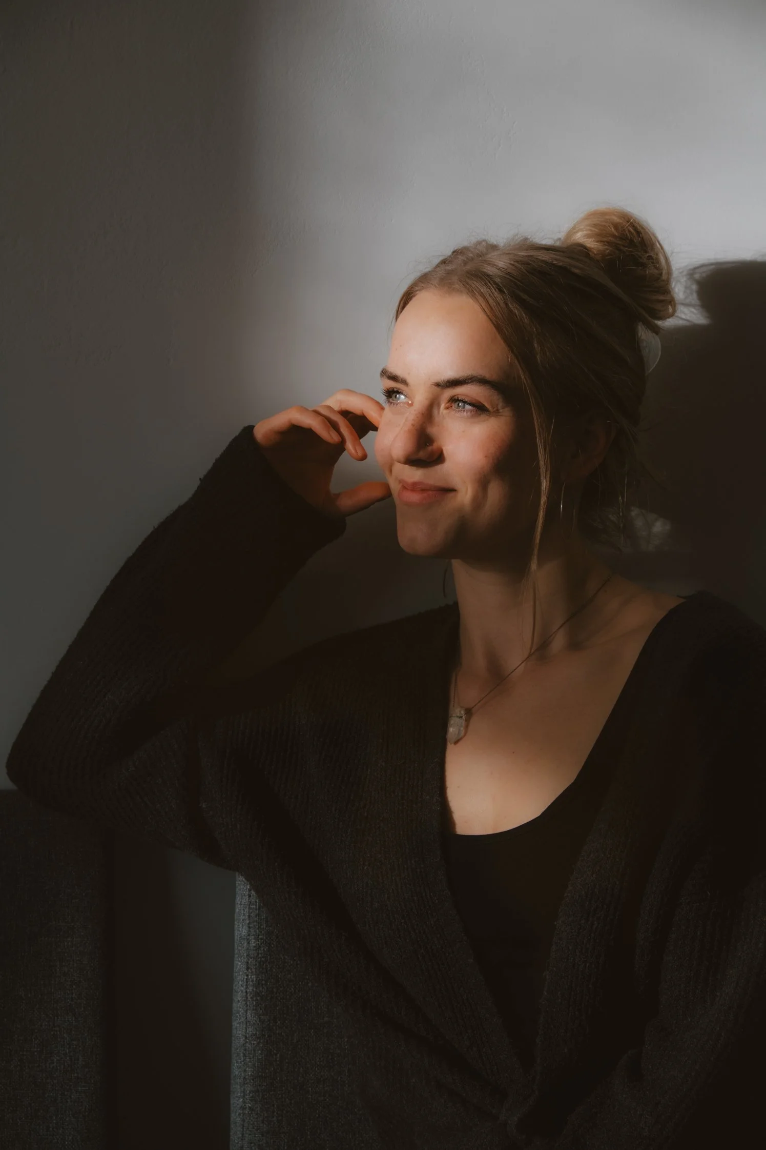 A young woman with blonde hair in a bun, wearing a black top and cardigan, sitting against a plain wall with soft lighting highlighting her face, smiling slightly and touching her face with her right hand.