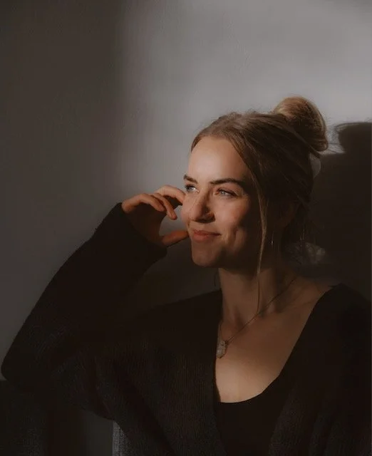 A young woman with a messy bun hairstyle, wearing a black top, smiling softly, with sunlight casting shadows on her face and background.