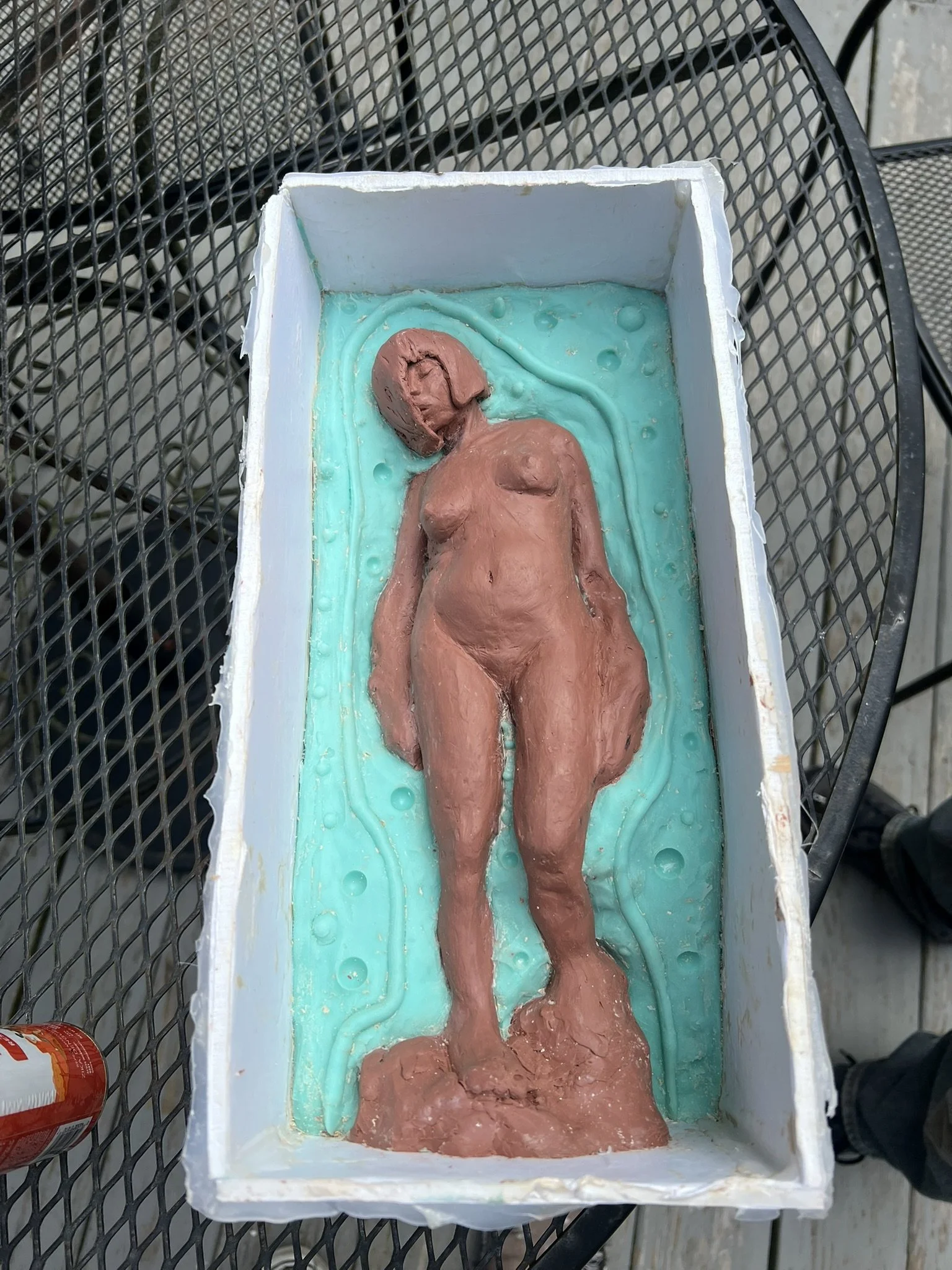 A sculpture of a nude woman lying on a light blue bed, with her face turned to the side, contained in a rectangular box.