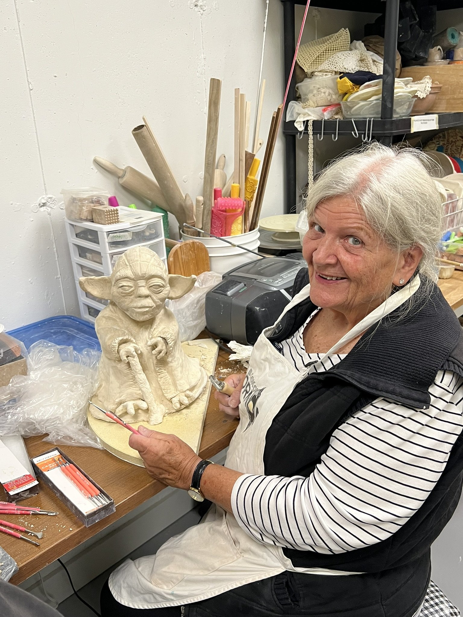 An elderly woman with gray hair, wearing a striped shirt and apron, sculpting a ceramic figure of Baby Yoda in a pottery studio.