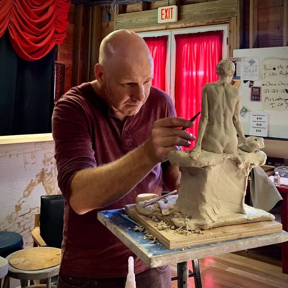 An artist sculpting a clay figure of a seated woman in a workshop with red curtains and wooden walls.