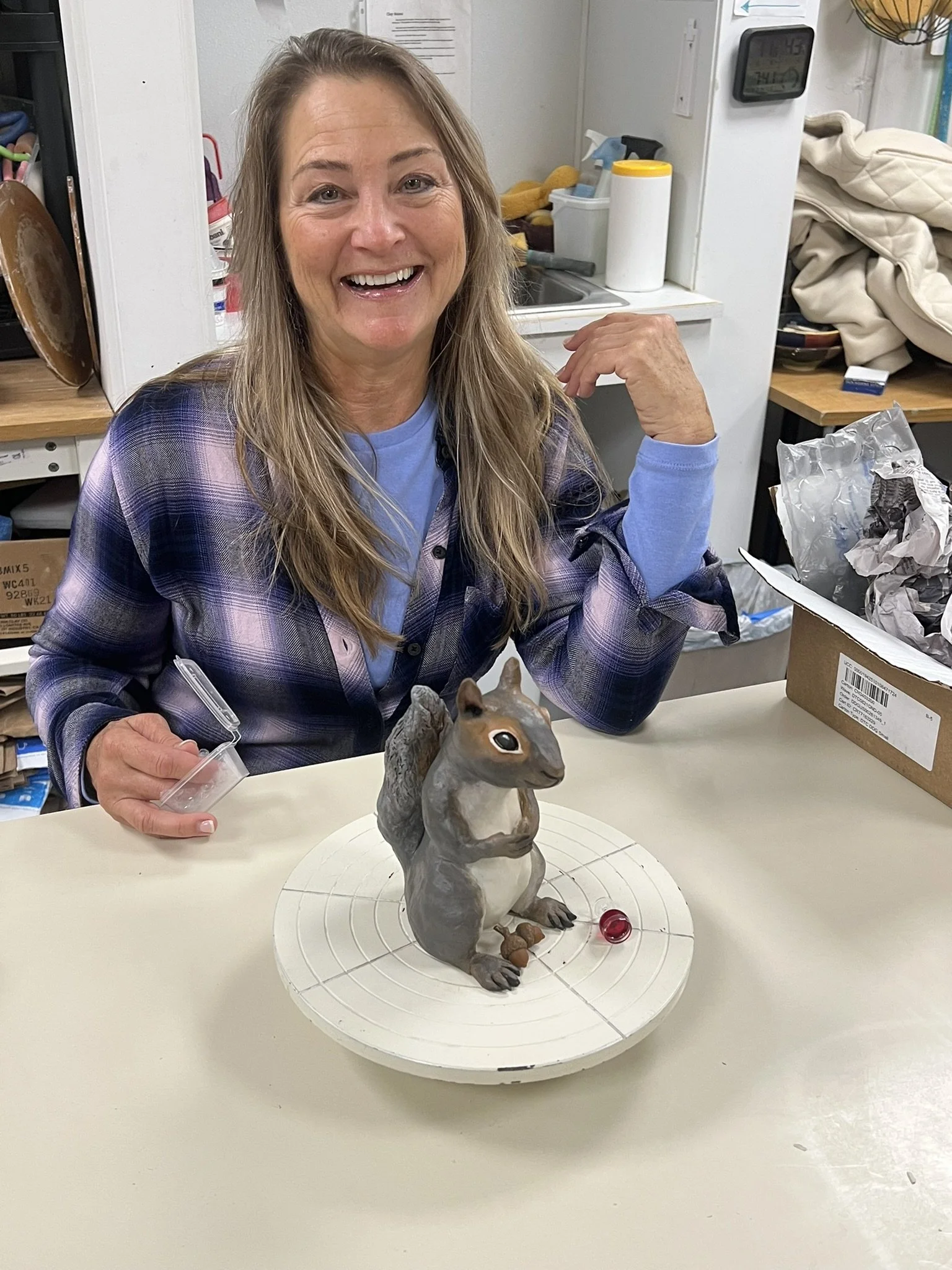 A woman sitting at a table next to a squirrel figurine with a large tail, with a small red object and brown acorns on the base, in a room with kitchen supplies and boxes.