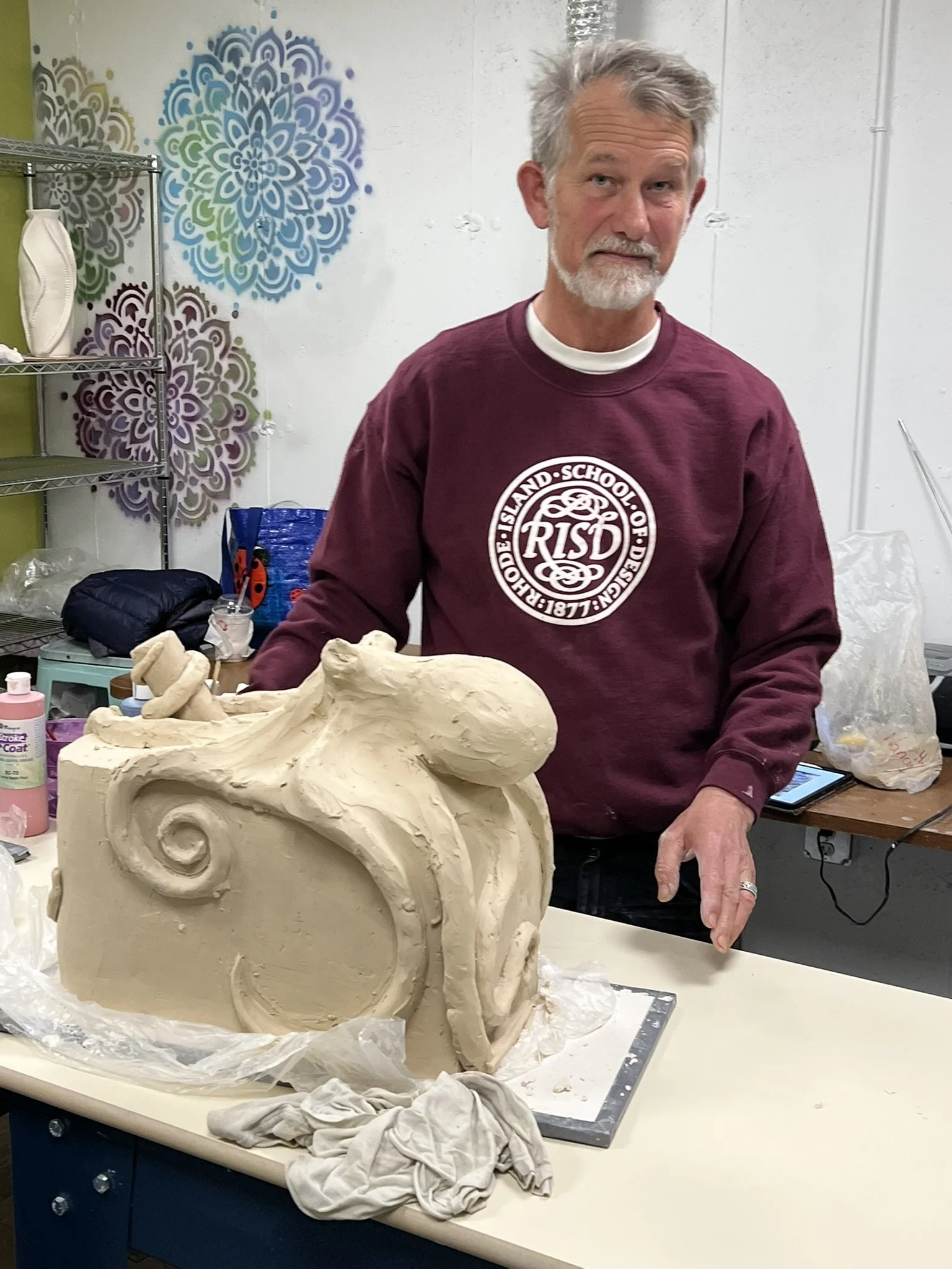 A man with gray hair and a beard wearing a maroon sweatshirt stands behind a large, unfinished clay sculpture on a table.