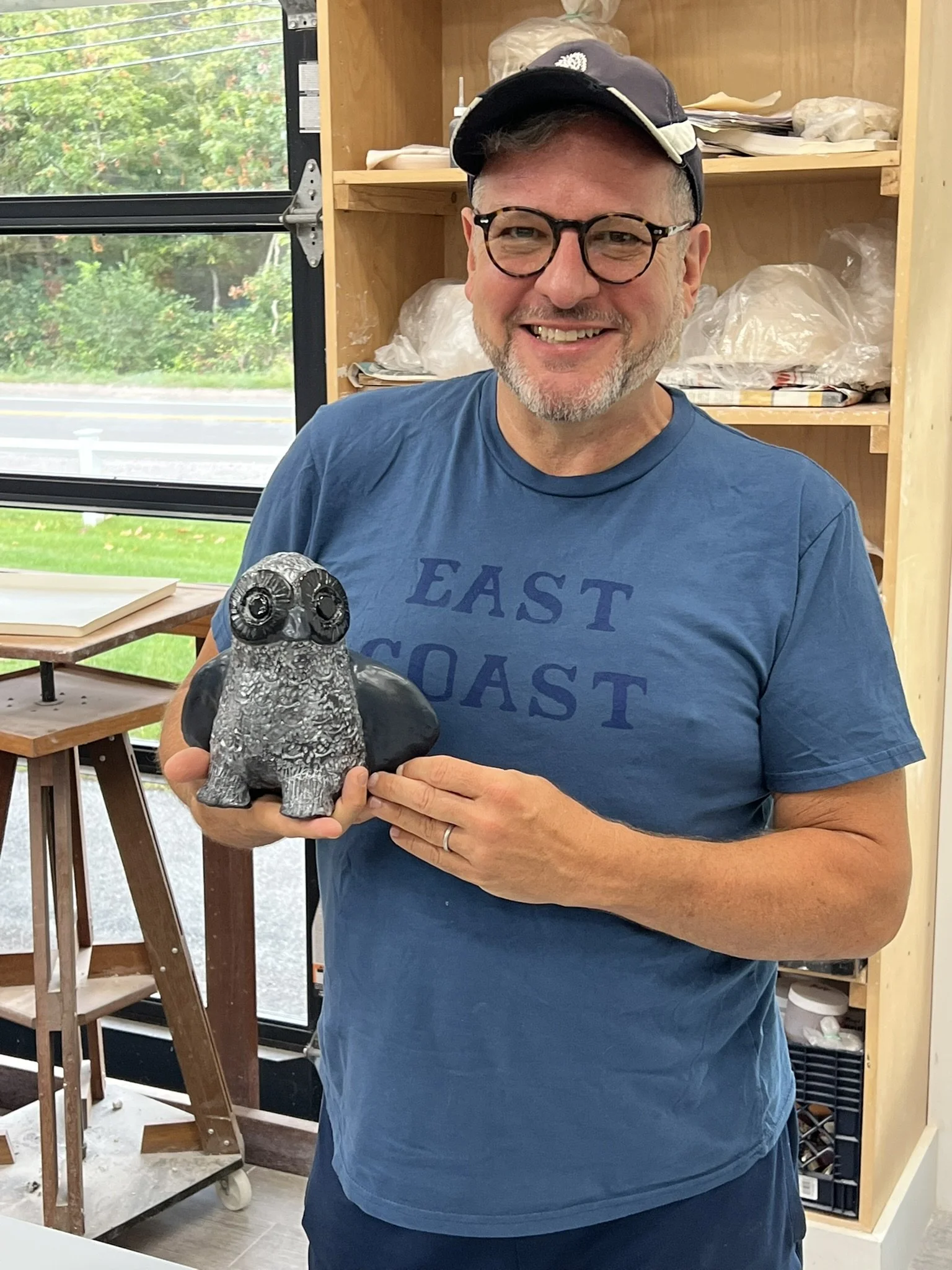 A man wearing glasses, a cap, and a blue "EAST COAST" t-shirt is smiling and holding a ceramic figurine of a bird with black wings in his right hand, inside a workshop.
