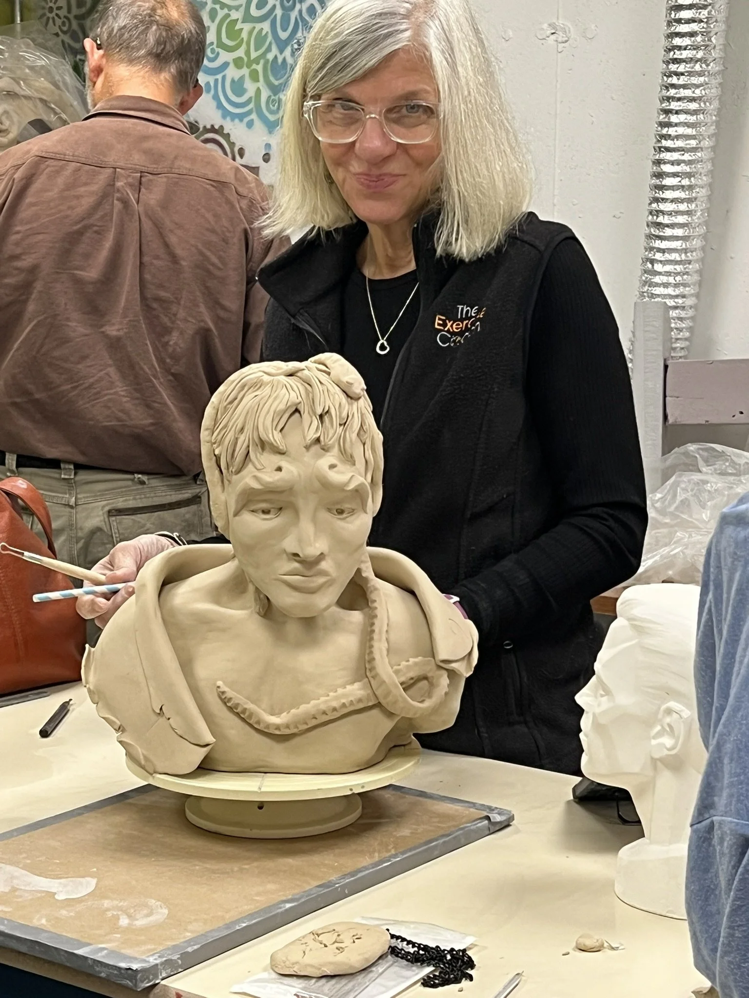 A woman with blonde hair, glasses, and a black vest is standing behind a clay sculpture of a person's head and upper torso. She is in an art studio or classroom, with other artworks and people in the background.