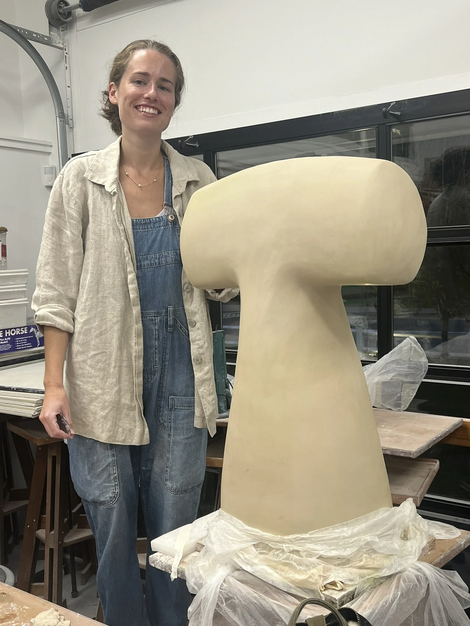A woman in a beige shirt and denim overalls standing next to a large ceramic or plaster sculpture that is in the shape of a mushroom.