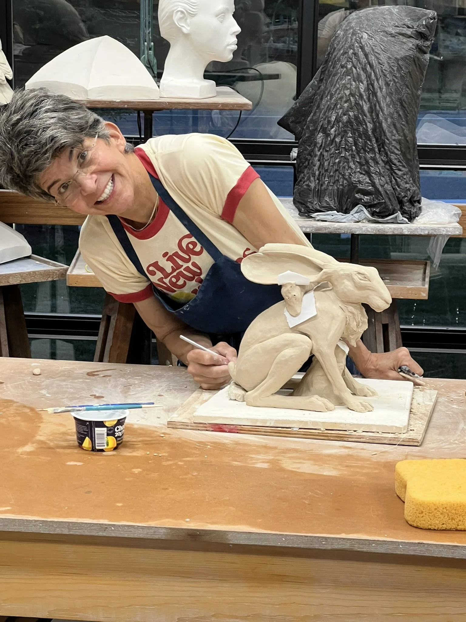 A woman smiling and working on a ceramic sculpture of a rabbit and a hare, with various art supplies and sculptures in the background.