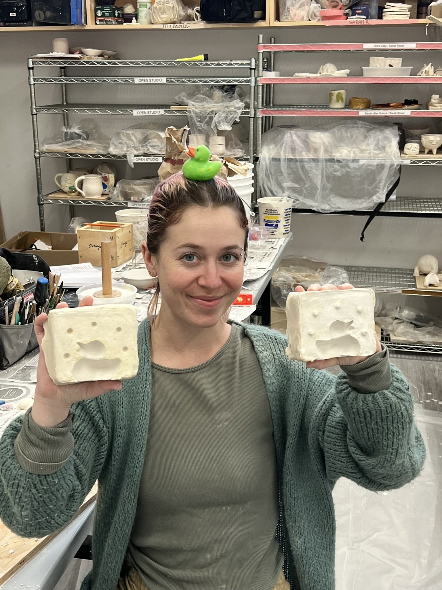 A woman in an art studio holding two blocks of cheese or soap with carved images of a sleeping bear. She wears a green cardigan and a small rubber duck on her head. Behind her are shelves with art supplies and tools.