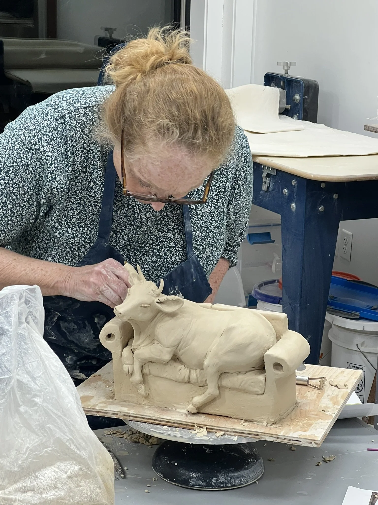 A woman with glasses and curly hair tied up, wearing a patterned shirt and apron, shaping a detailed clay sculpture of a reclining cow on a work table in a studio.