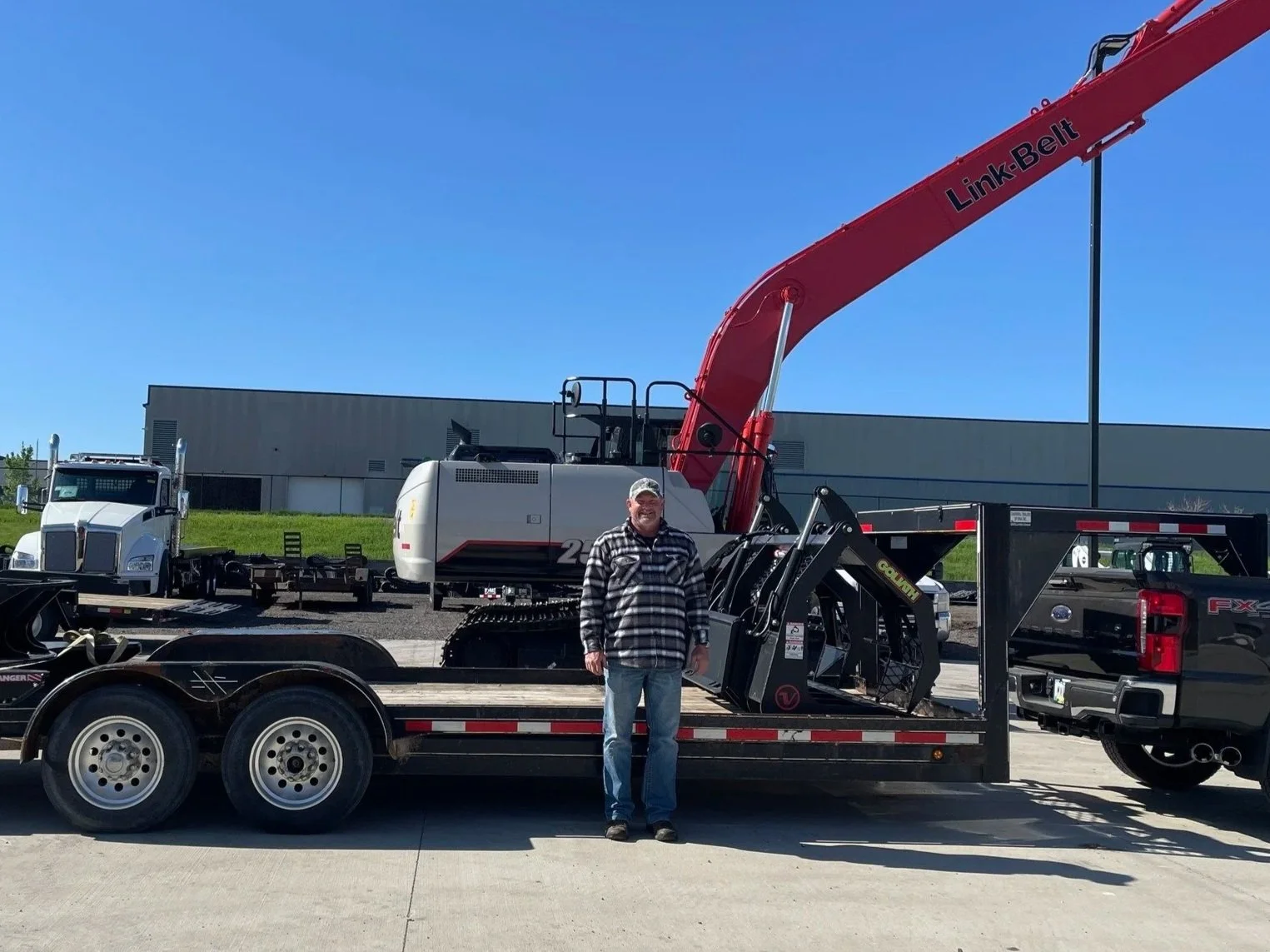 A man standing on a flatbed trailer with a large excavator and a crane in the background. The man is wearing a black and white plaid jacket and a baseball cap. The excavator has a long red arm labeled "Link-Belt" and tracks instead of wheels.