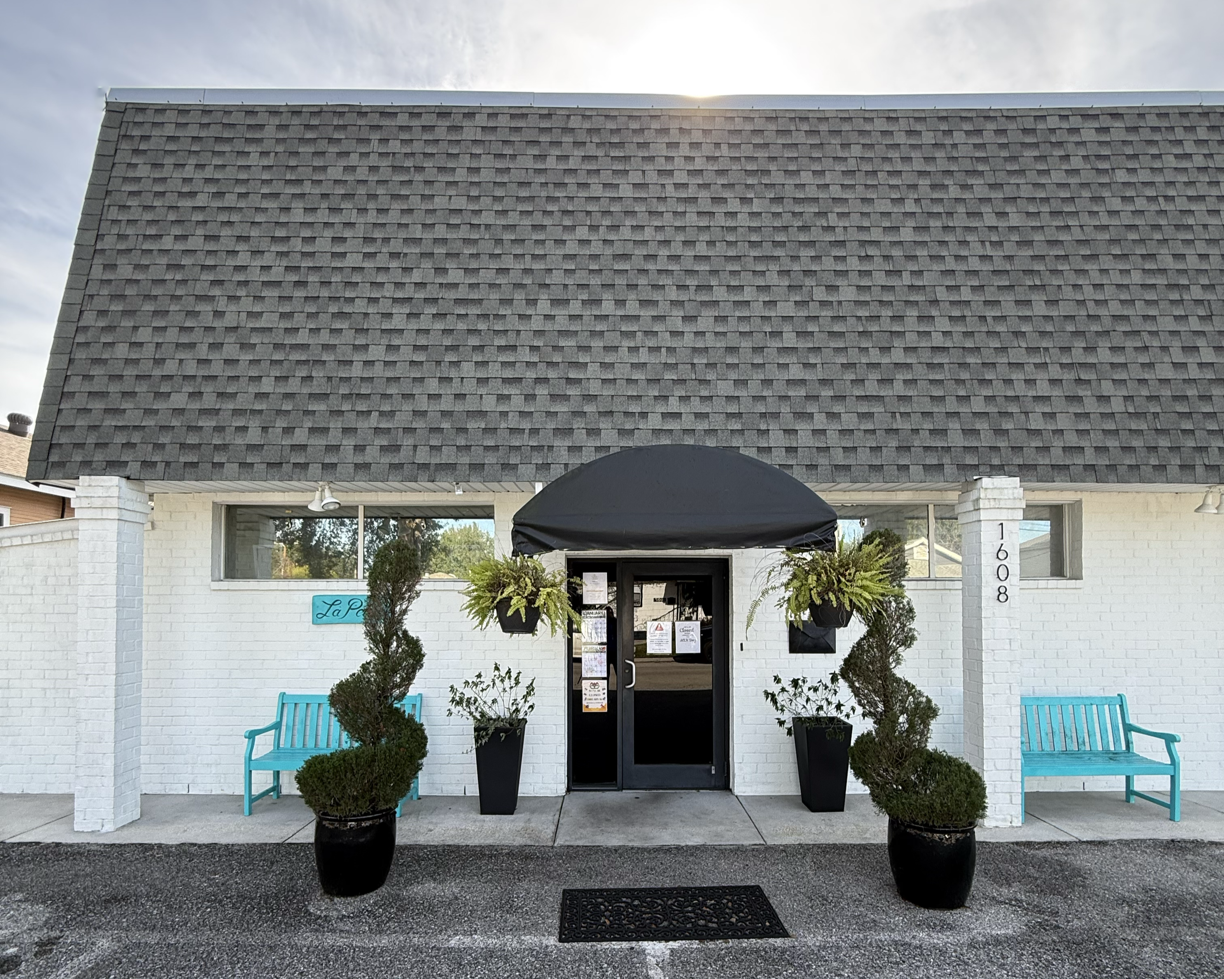 Exterior of a white brick building with a gray shingled roof, black awning over the entrance, and two blue benches on either side of the door. Potted plants and greenery decorate the entrance.