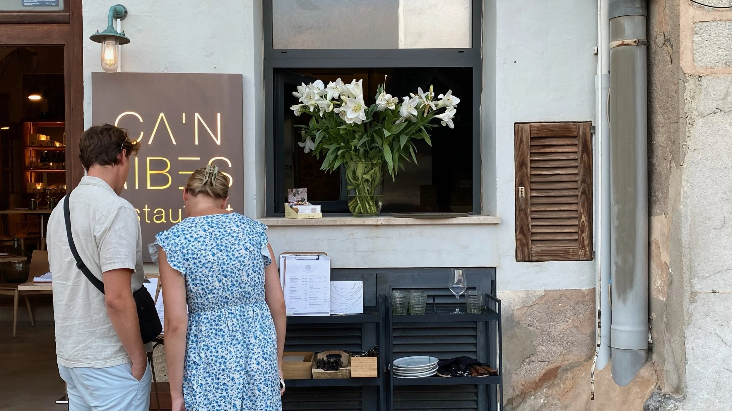Dos personas viendo un menú en la entrada de un restaurante con un cartel que dice 'CAN RIBES' y un jarrón con flores blancas en la ventana.