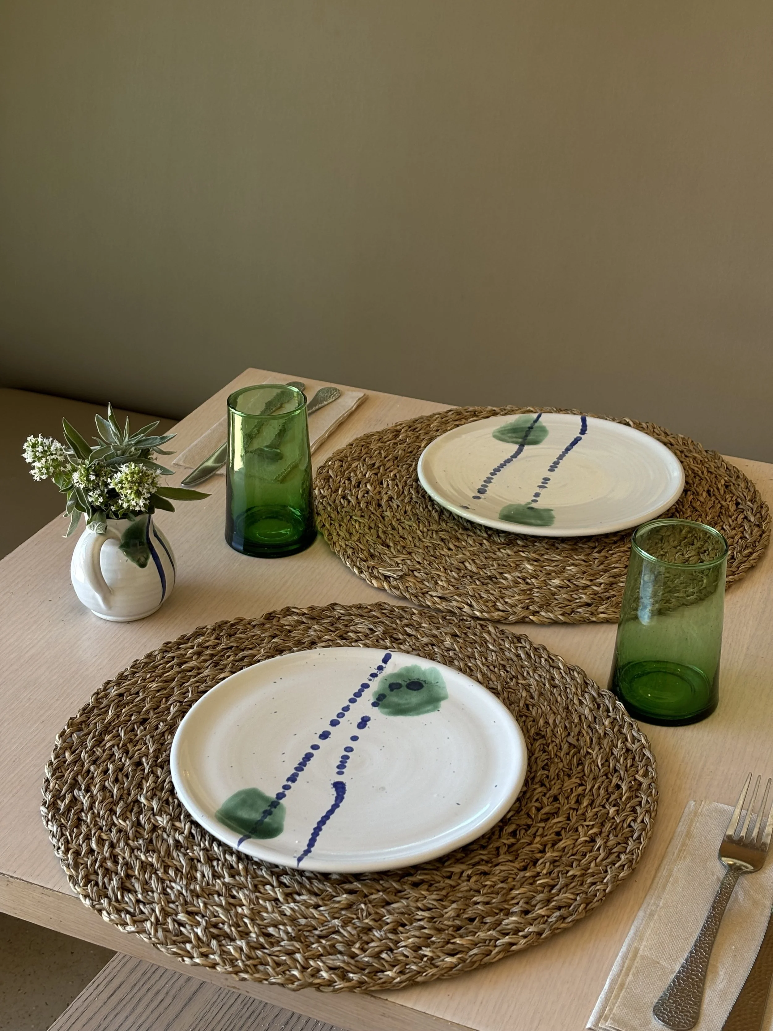 Mesa preparada con platos blancos decorados con puntos y líneas verdes y azules en platos grandes, con vasos verdes y un pequeño florero con flores en una mesa de madera del restaurante Ca'n Ribes.