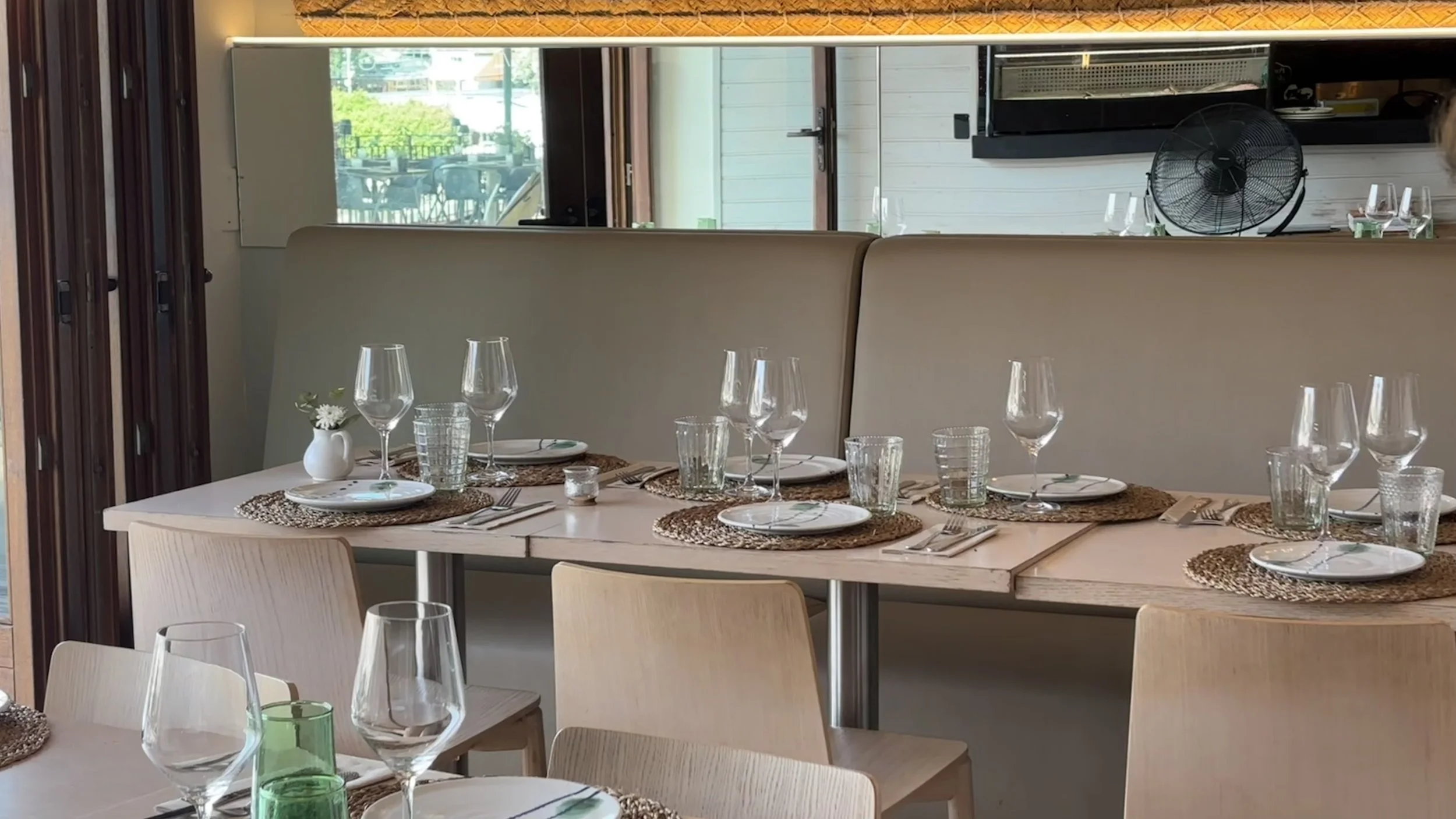 Mesa situada en el salón de abajo preparada para comer con platos, copas de vino, vasos, y cubiertos en un restaurante con un sofá y la puerta totalmente abierta que deja entrar luz natural y se ven las vistas del Puerto de Sóller.