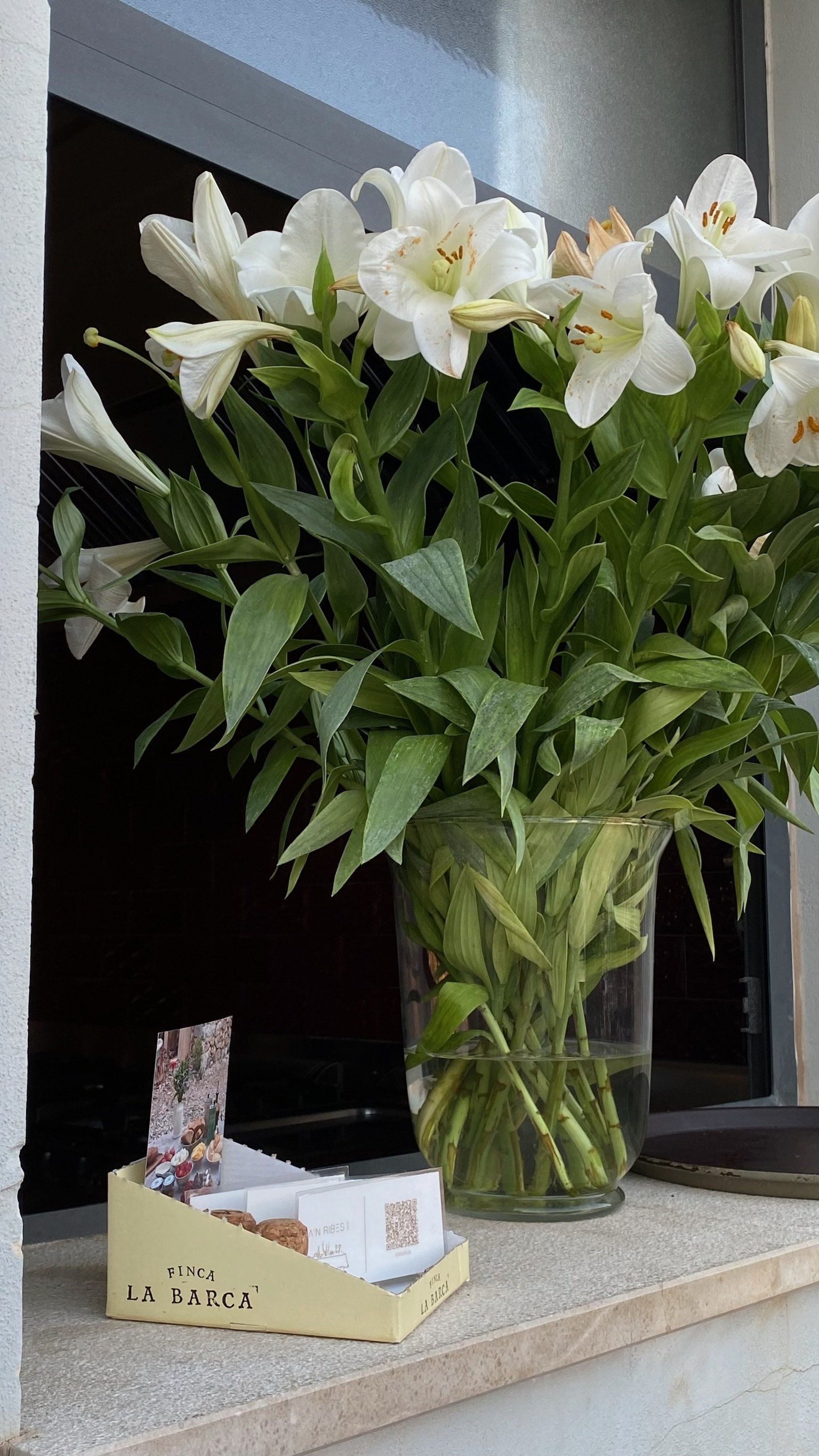 Un jarrón con flores blancas de lirio en un jarrón junto a una caja pequeña amarilla de la marca La Barca.