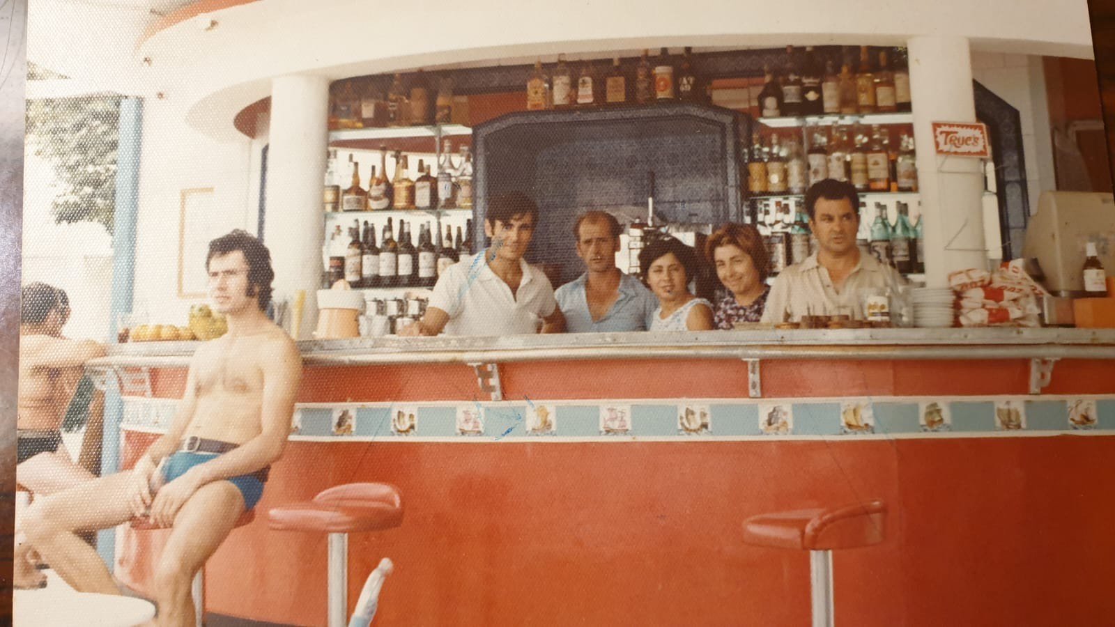Una escena de un bar con varios clientes, uno de ellos en bañador, en un ambiente con decoración vintage y visible estantería con botellas y utensilios. Era el antiguo quiosco del padre de los dueños.