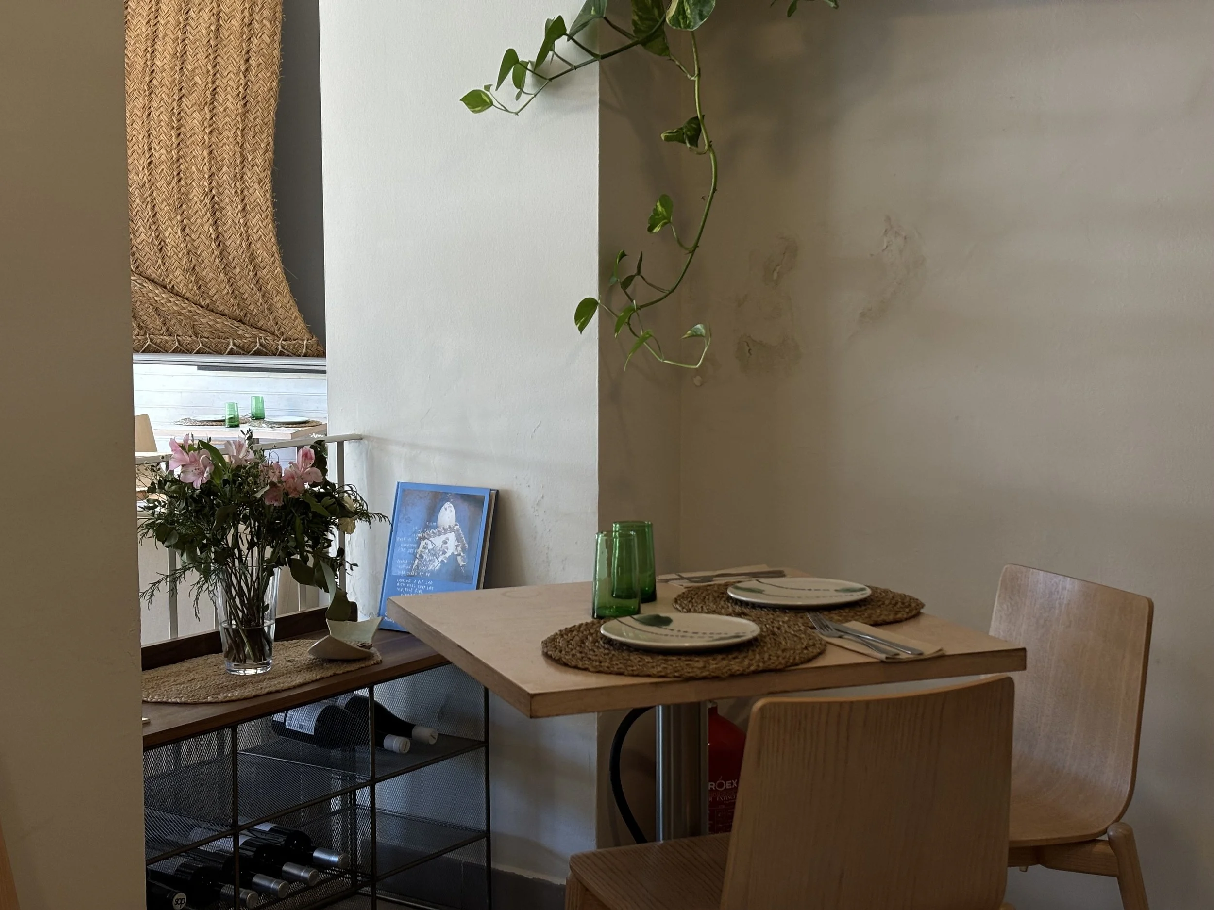 Mesa de comedor con platos, vasos verdes y manteles de yute, cerca de una pared blanca con plantas colgantes, decorada con un ramo de flores y un libro de Apartamento sobre la casa de Dalí, en el salón del restaurante.