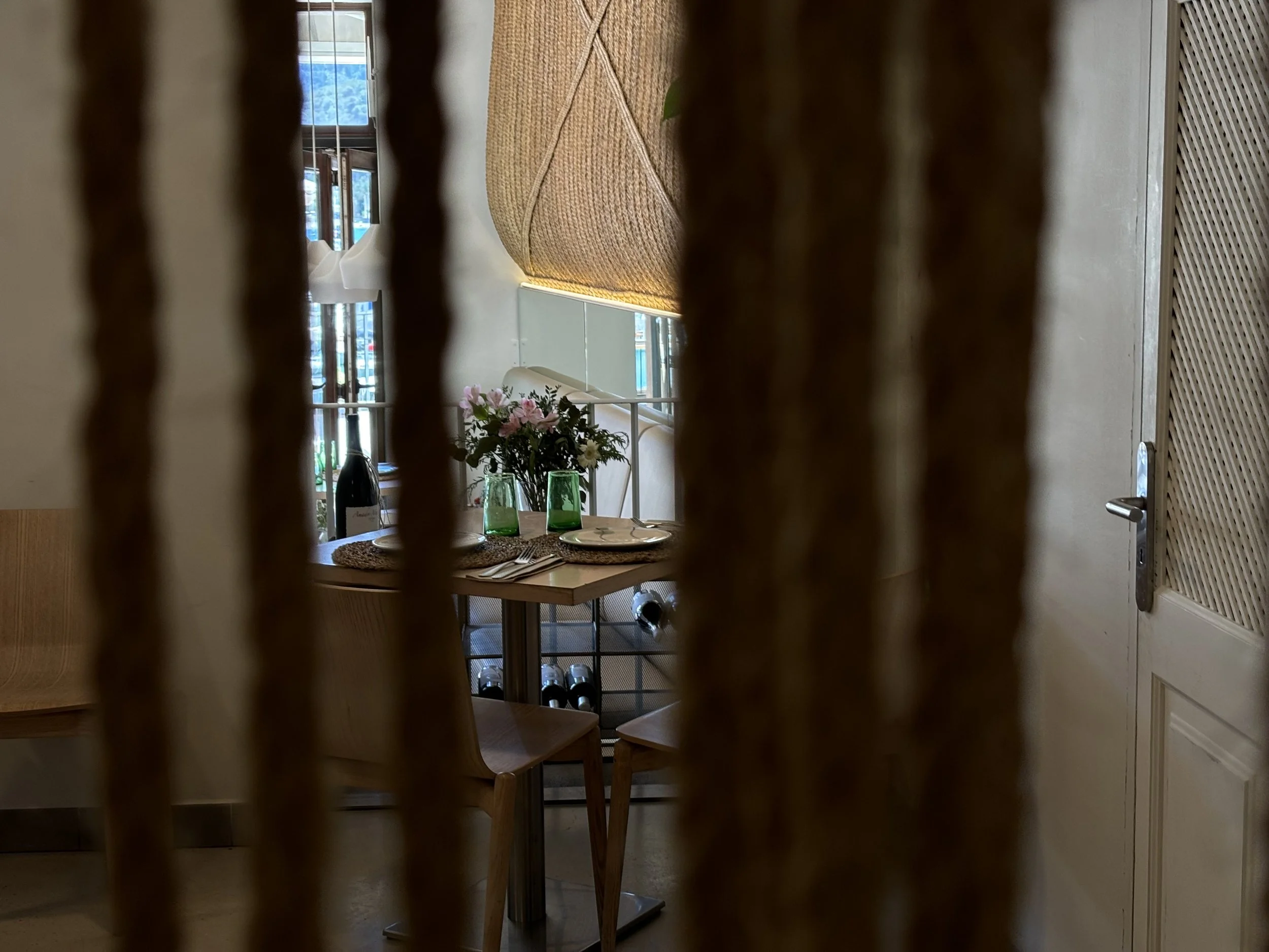 Vista de una mesa de comedor vista a través de unas cortinas de cuerda, con platos, vasos y flores decorativas.