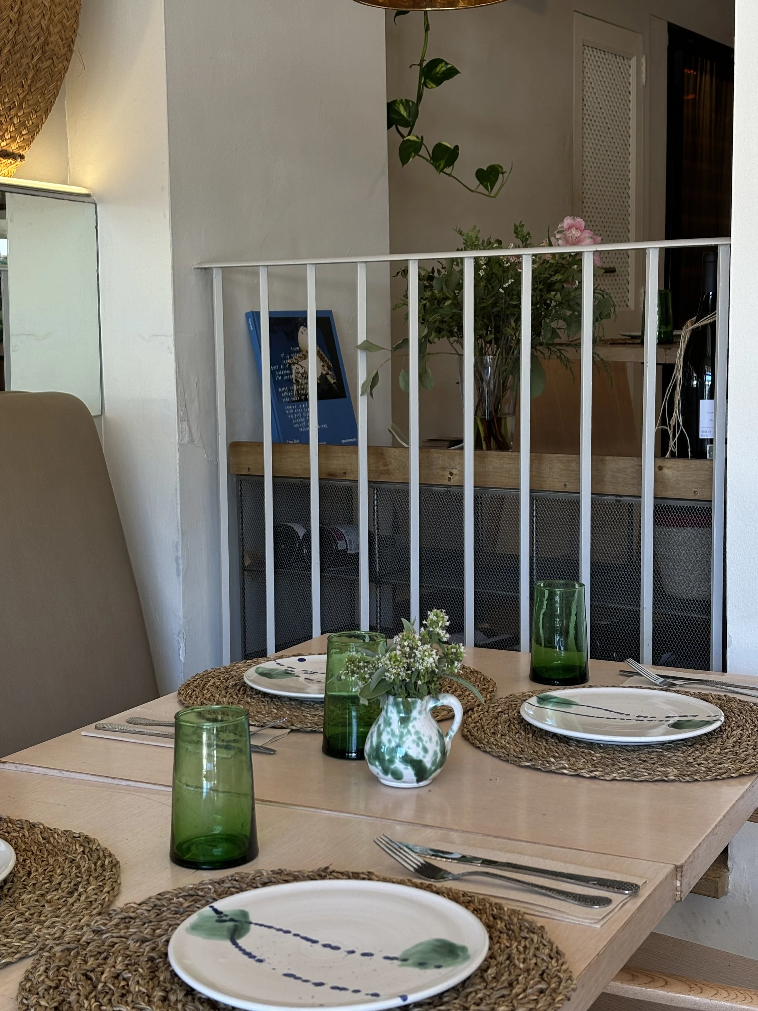 Mesa puesta para comer con platos, vasos de vidrio verde, cubiertos y un jarrón con flores en el centro en un ambiente de comedor interior.